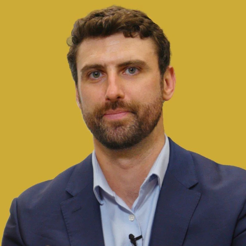 A man with brown hair and a beard wearing a blue suit jacket and light blue shirt against a yellow background.