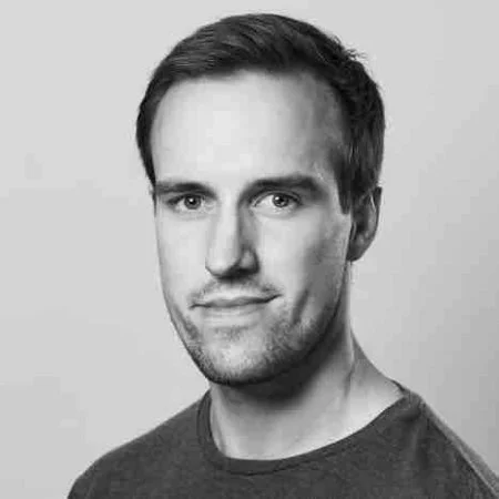 A black-and-white headshot of a young man with short dark hair, light facial hair, and wearing a round-neck shirt, looking at the camera with a slight smile.
