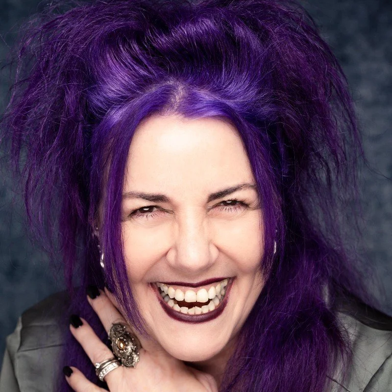 Woman with vibrant purple hair smiling widely, wearing dark lipstick, rings, and earrings, against a dark background.