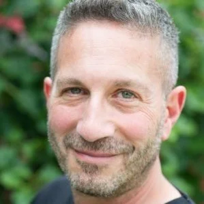 Close-up of a smiling middle-aged man with short gray hair and a beard, outdoors with greenery in the background.
