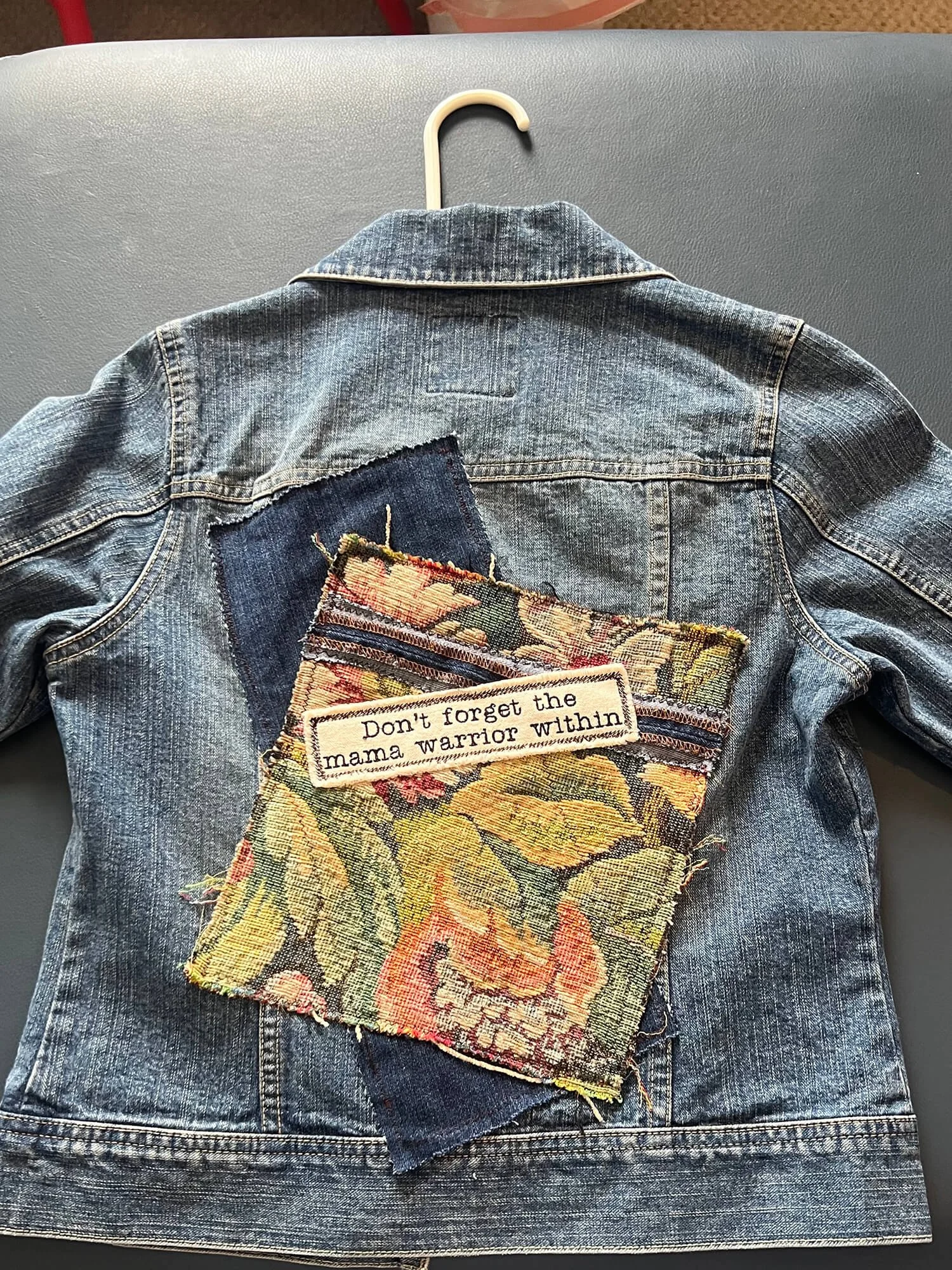 A denim jacket hanging on a grey surface with a fabric patch on the back that reads, "Don't forget the mama warrior within."