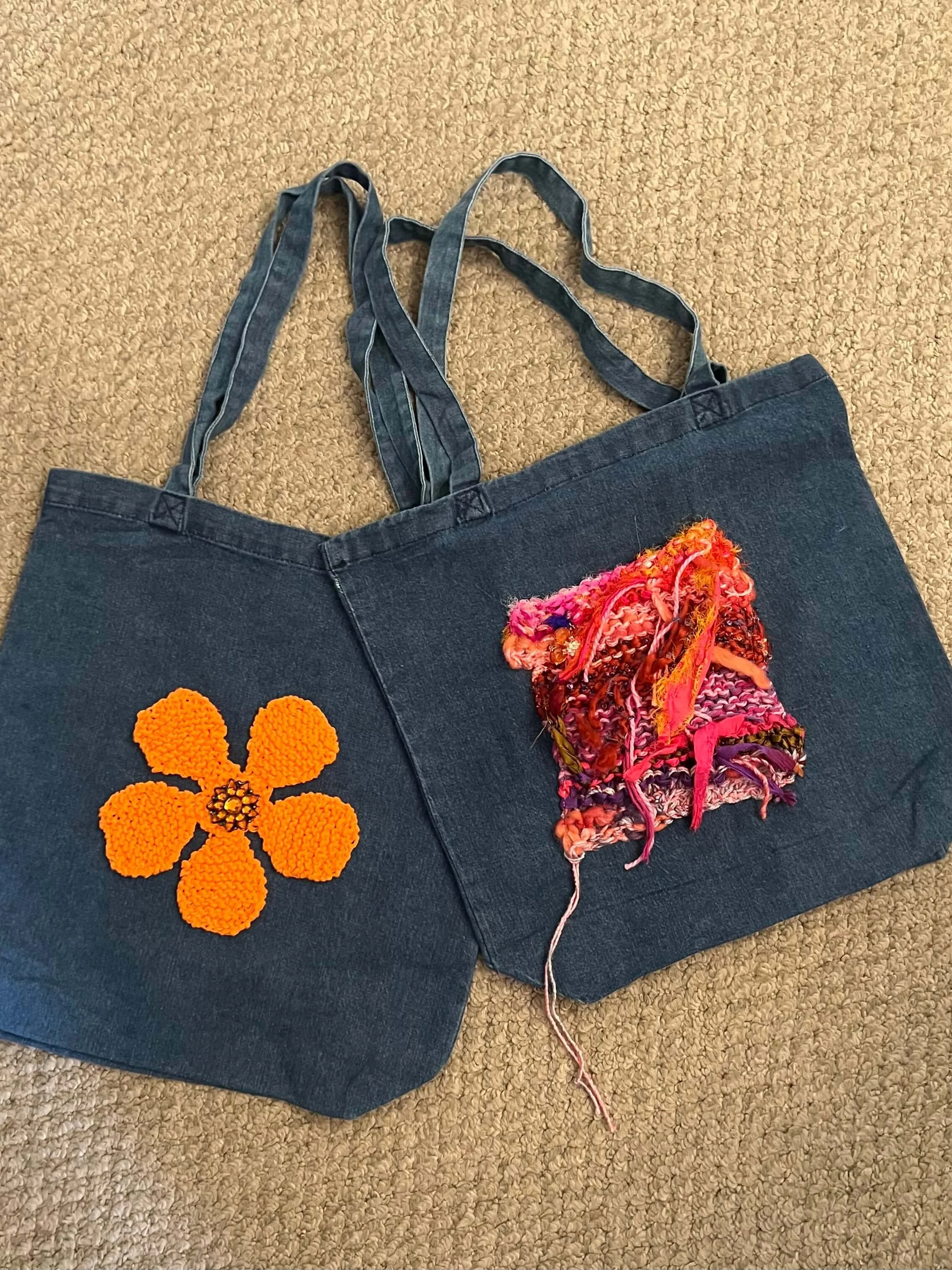 A dark blue denim tote bag with two colorful fabric patches, one being a bright orange flower and the other a multicolored textured square, placed on a beige carpet.