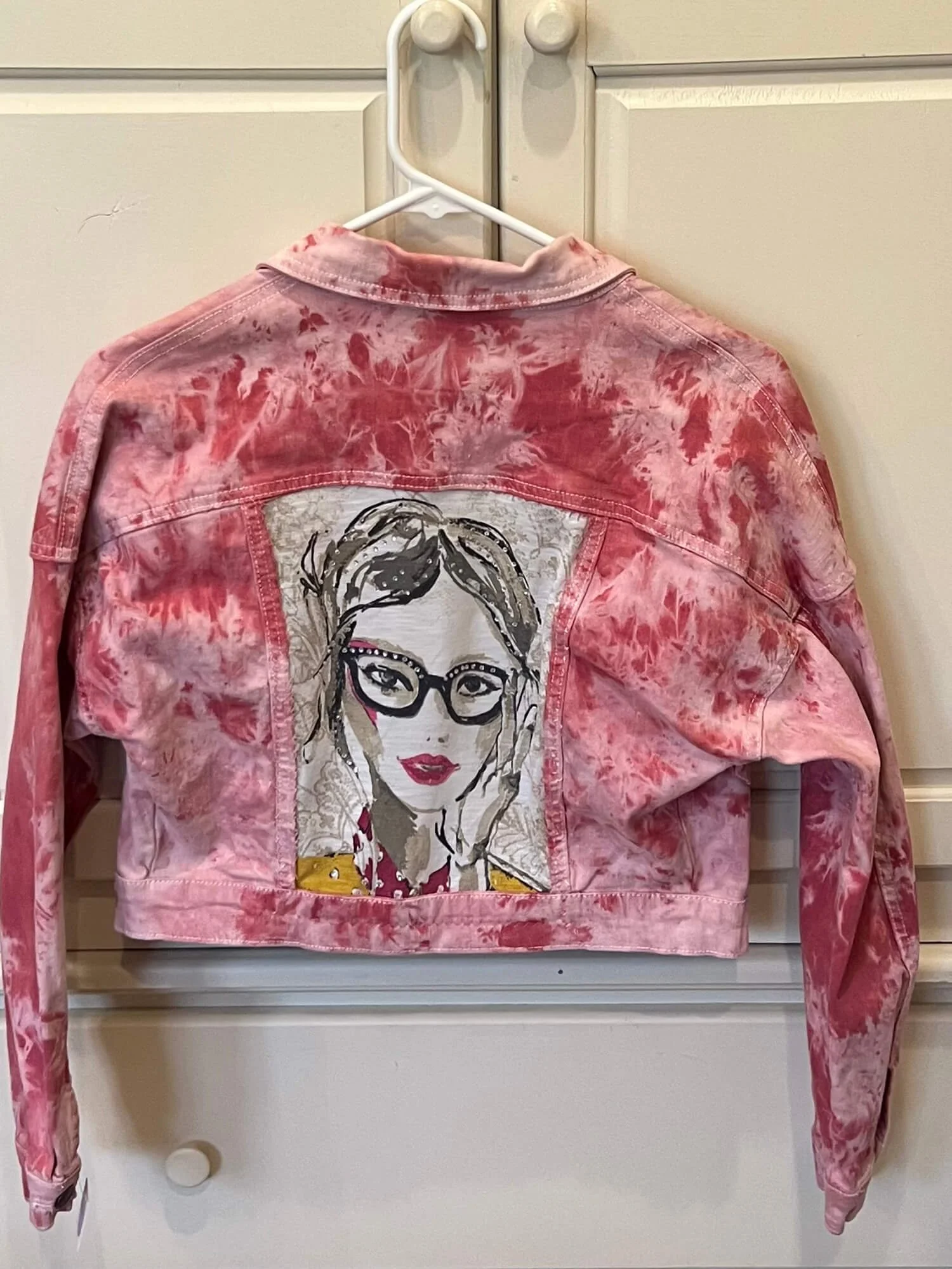 Pink tie-dye denim jacket with a portrait of a woman with glasses and red lipstick embroidered on the back.