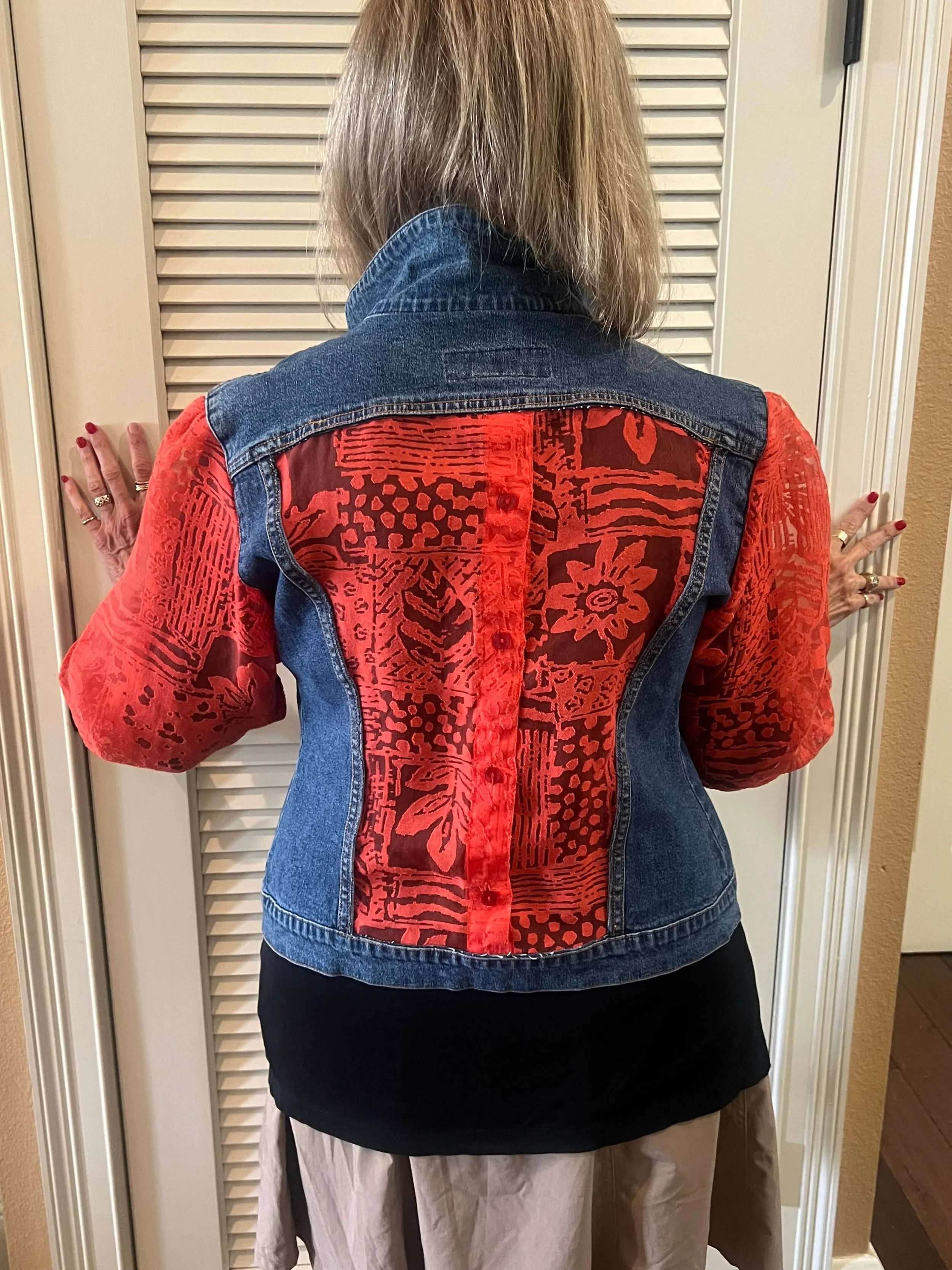 Back view of a woman with shoulder-length blonde hair wearing a denim vest with a red floral and abstract patterned back and red sleeves, standing in front of a beige closet door.