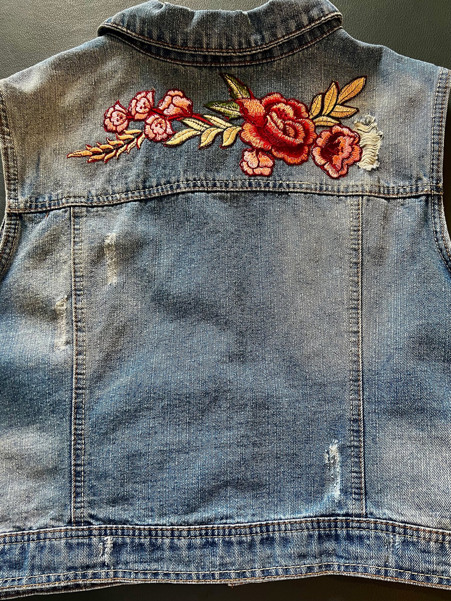 Back of a denim vest with embroidered red and pink flowers and green leaves.