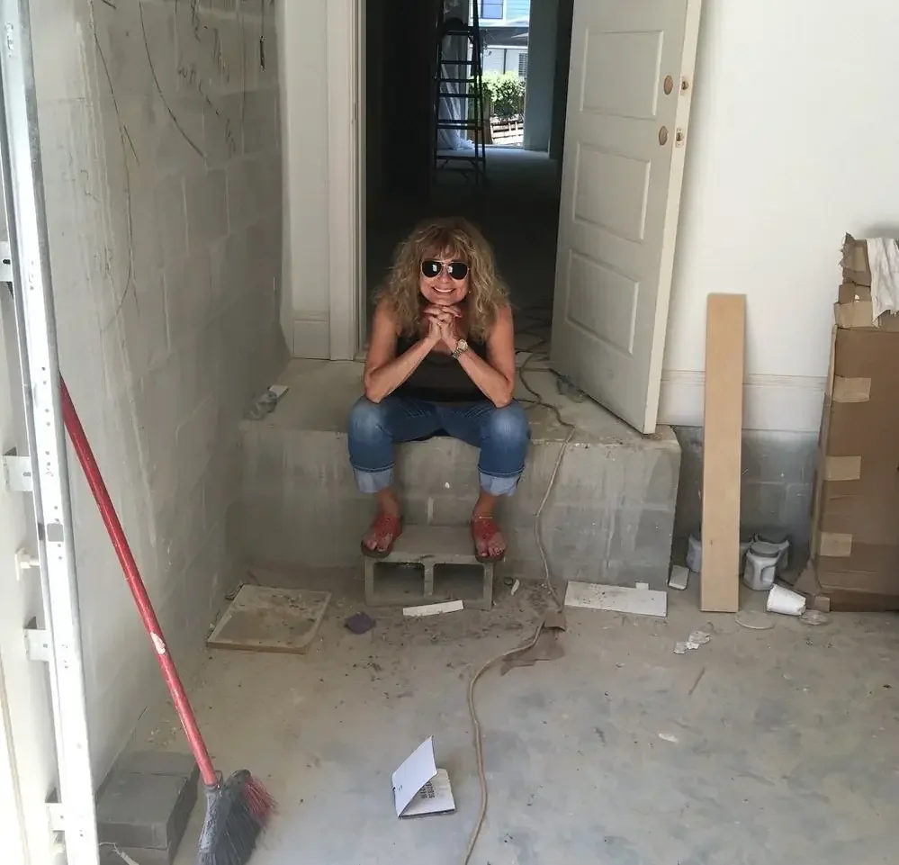 A woman with curly hair and sunglasses sitting on a small step inside a house under renovation, smiling with her chin resting on her hands.