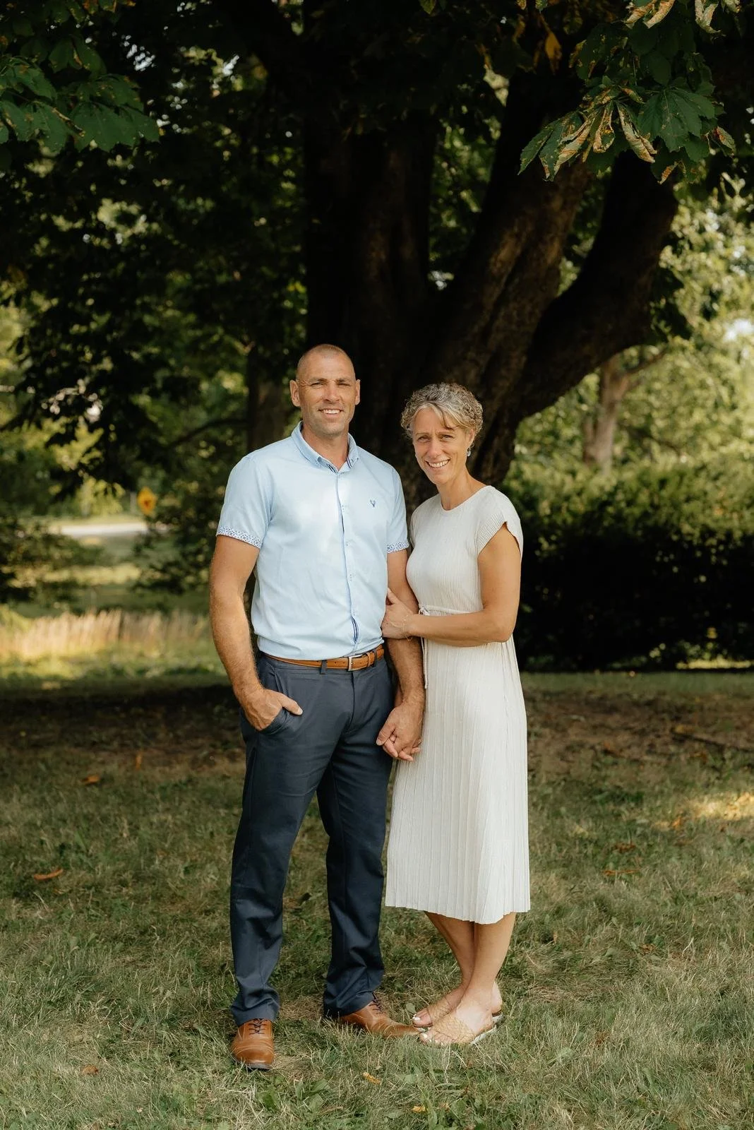 A middle-aged couple standing arm in arm outdoors in front of a large tree, smiling at the camera.