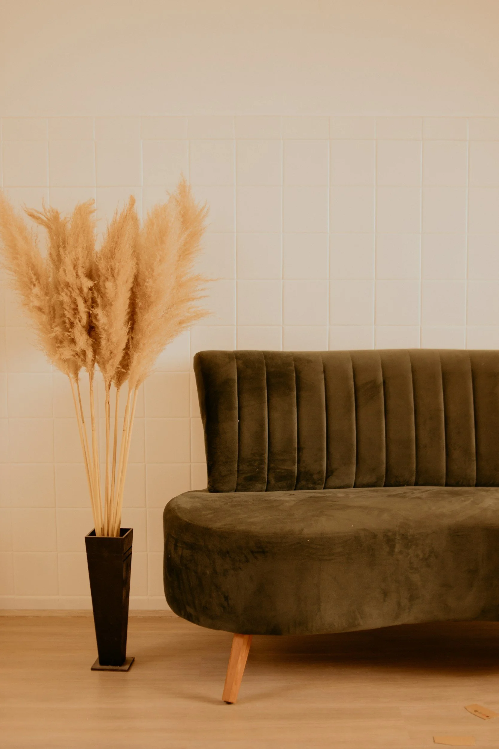 A dark green velvet sofa with wooden legs next to a tall black vase containing beige pampas grass, all placed against a white tiled wall and light wooden floor.