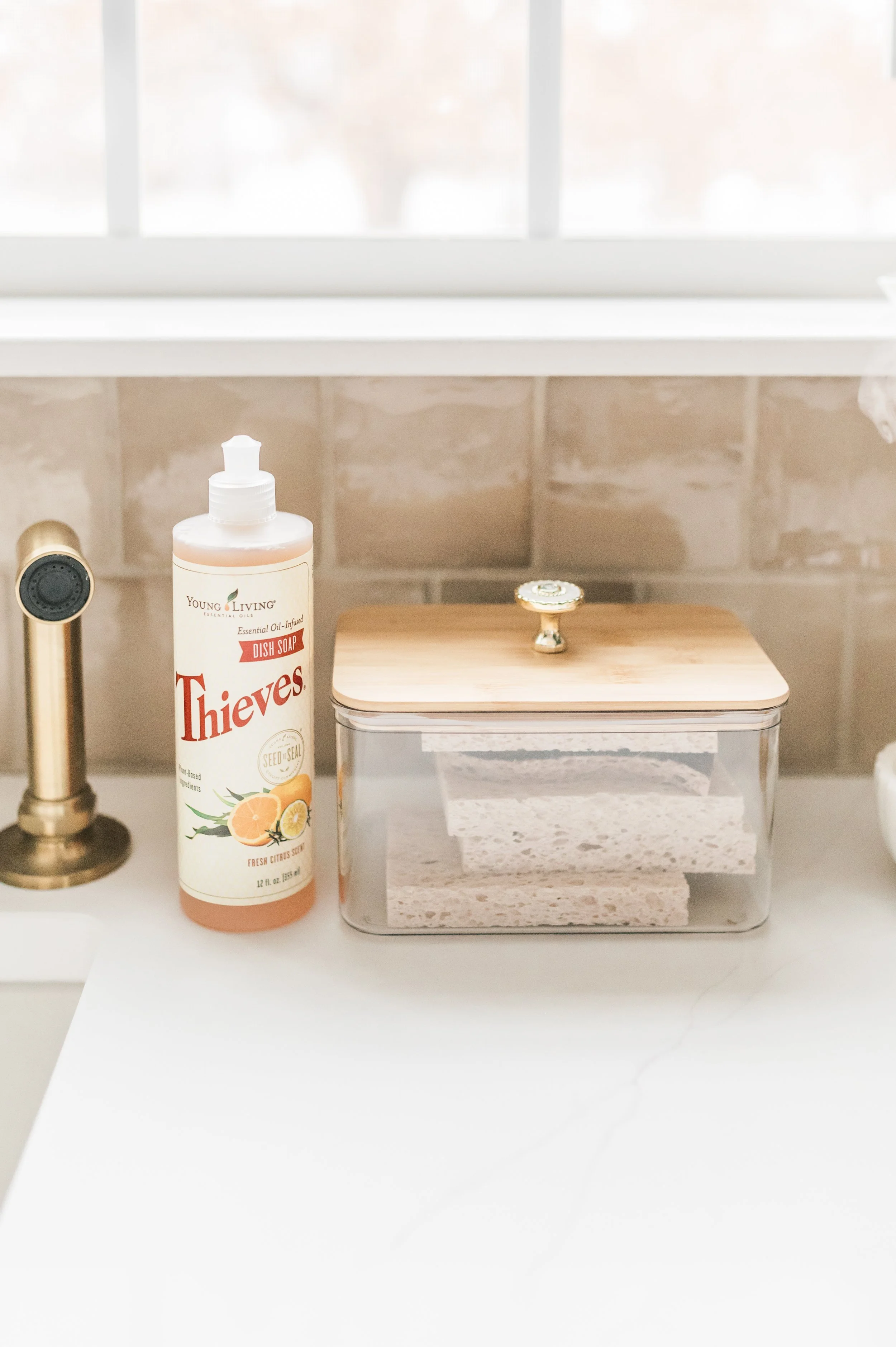 A kitchen countertop with a bottle of Young Living Thieves dish soap, a soap dispenser, and a container with a wooden lid filled with sponges. A window is in the background.