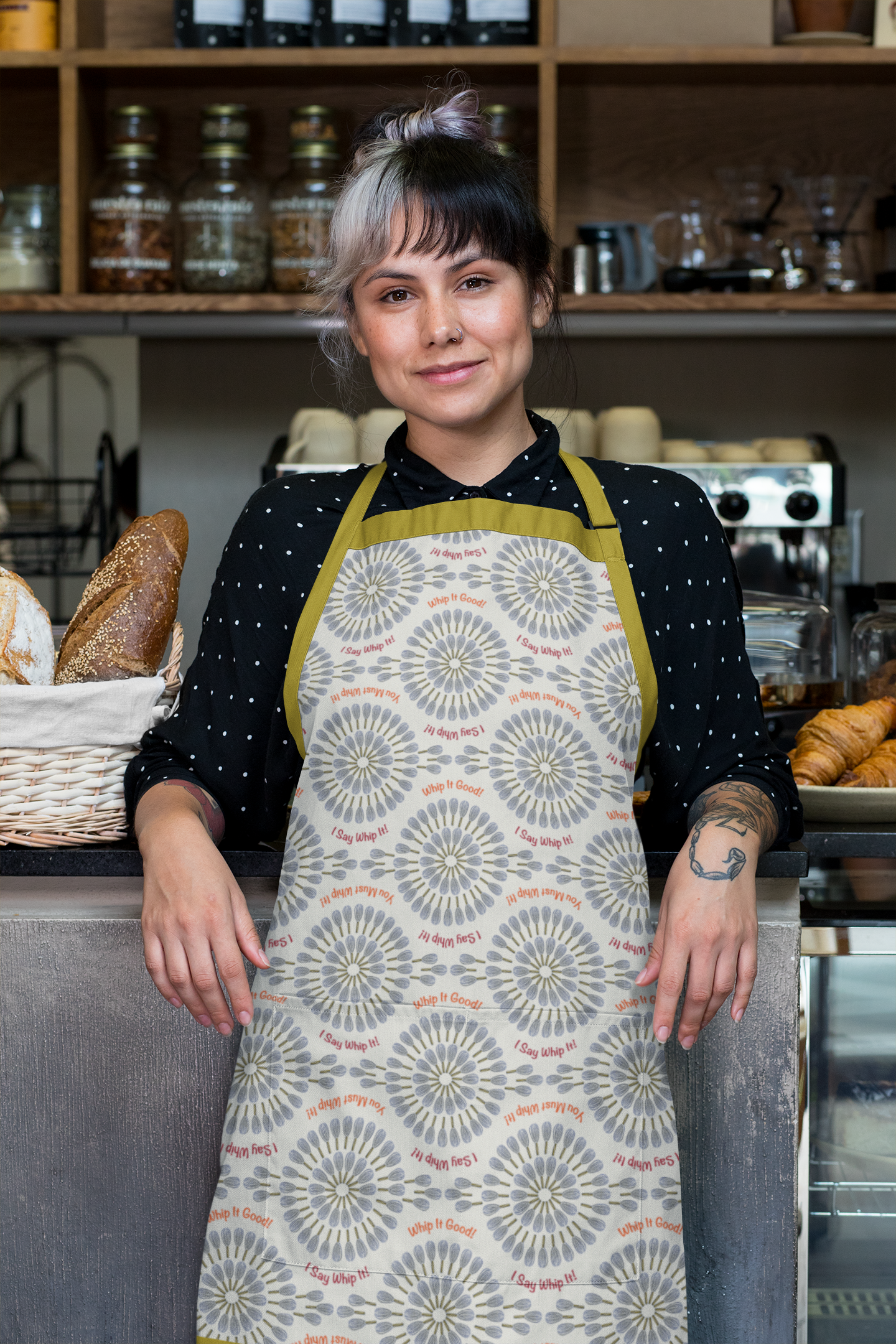 A woman with black and white hair, wearing a black polka dot shirt and a floral apron, stands behind a counter at a bakery, with bread and pastries in the background.