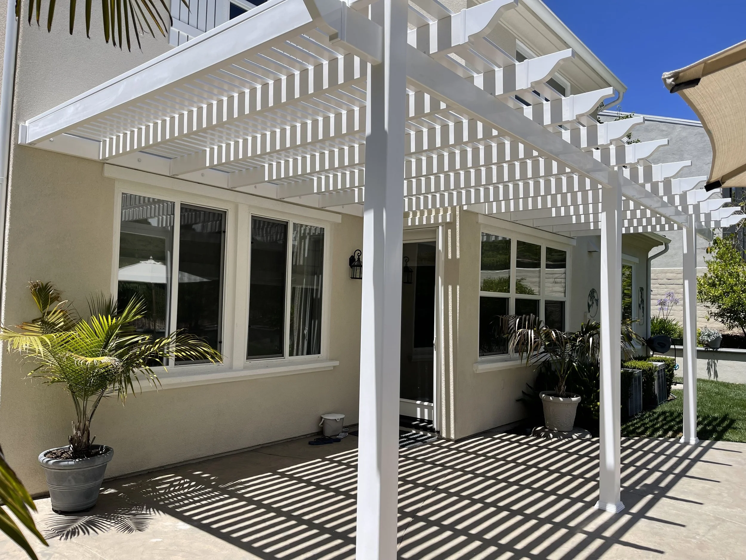 Backyard patio with white pergola casting striped shadows, featuring potted plants, windows, and a part of a house with a beige exterior and a sliding glass door.