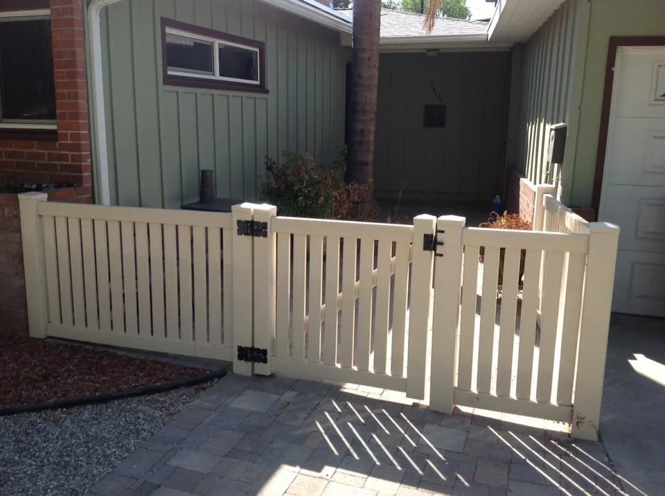 La Habra tan vinyl picket-style courtyard gate with black hinges, installed between brick and siding walls, leading to a front entry walkway, durable and stylish fencing for Orange County homes.