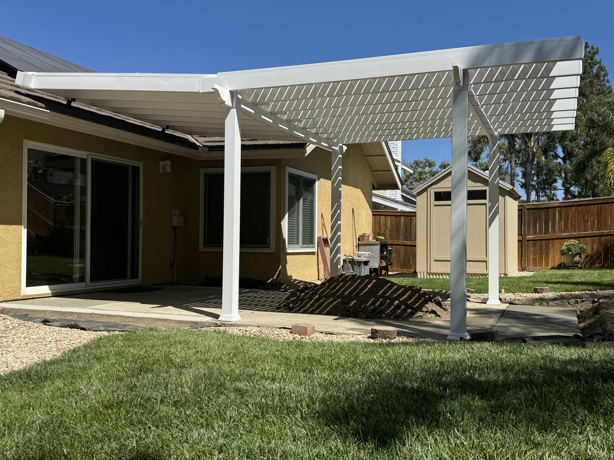 Aliso Viejo backyard patio with a white vinyl pergola, yellow house with sliding glass door, sliding window, small shed, fence and lush green grass, showcasing stylish vinyl pergolas, fencing and custom gates for Orange County homes.