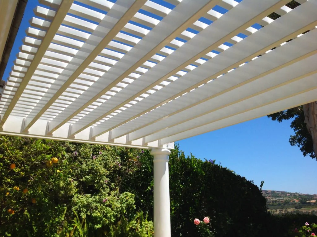 Laguna Hills white vinyl trellis or pergola with slats supported by a white column in a garden with green bushes and pink flowers under a clear blue sky, showcasing stylish vinyl patio covers and fences in Orange County.