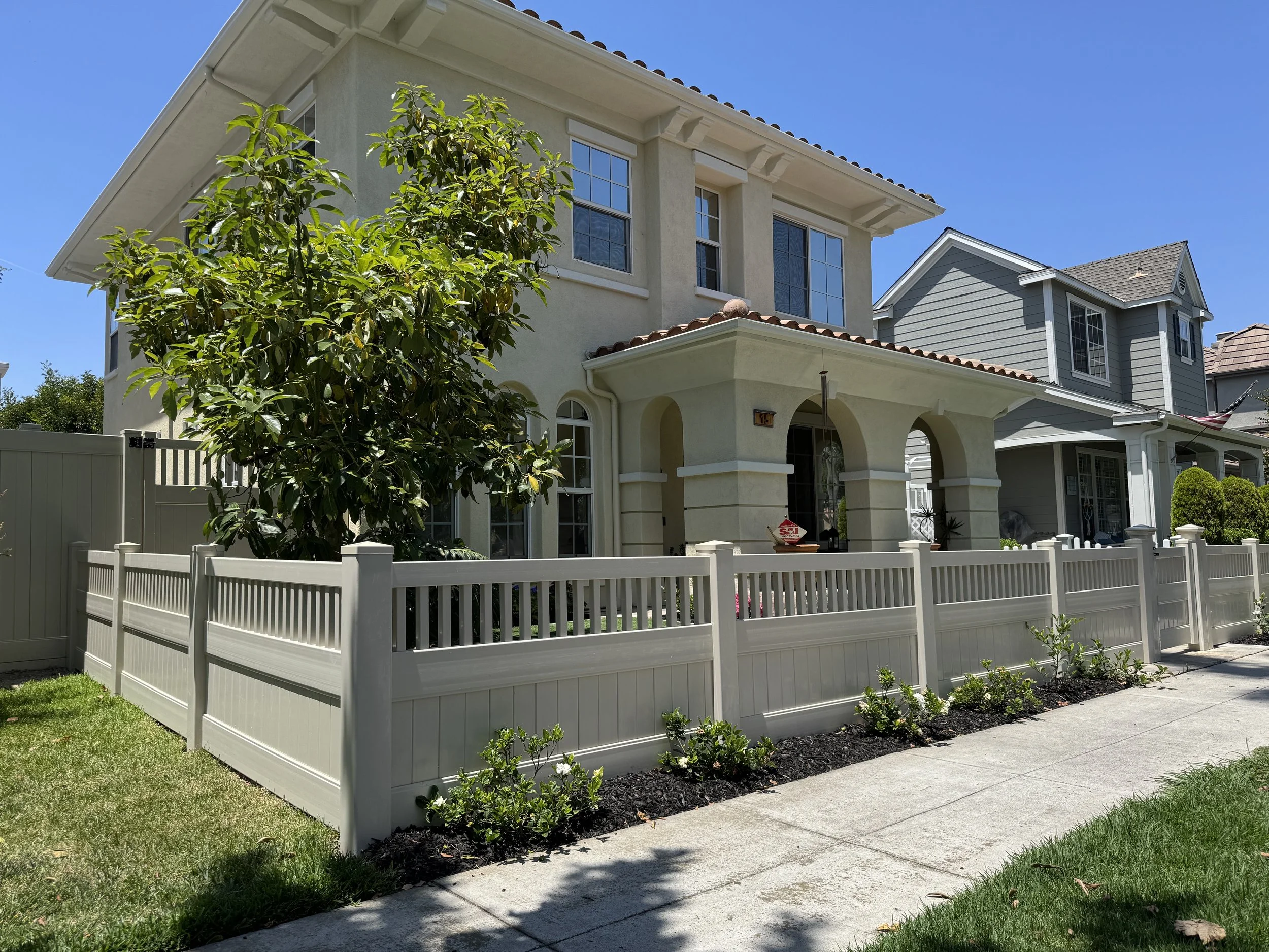 Villa Park two-story cream-colored house with front porch and white vinyl fence, large tree and manicured front yard under clear blue sky, showcasing premium vinyl fencing and custom gates for Orange County homes.