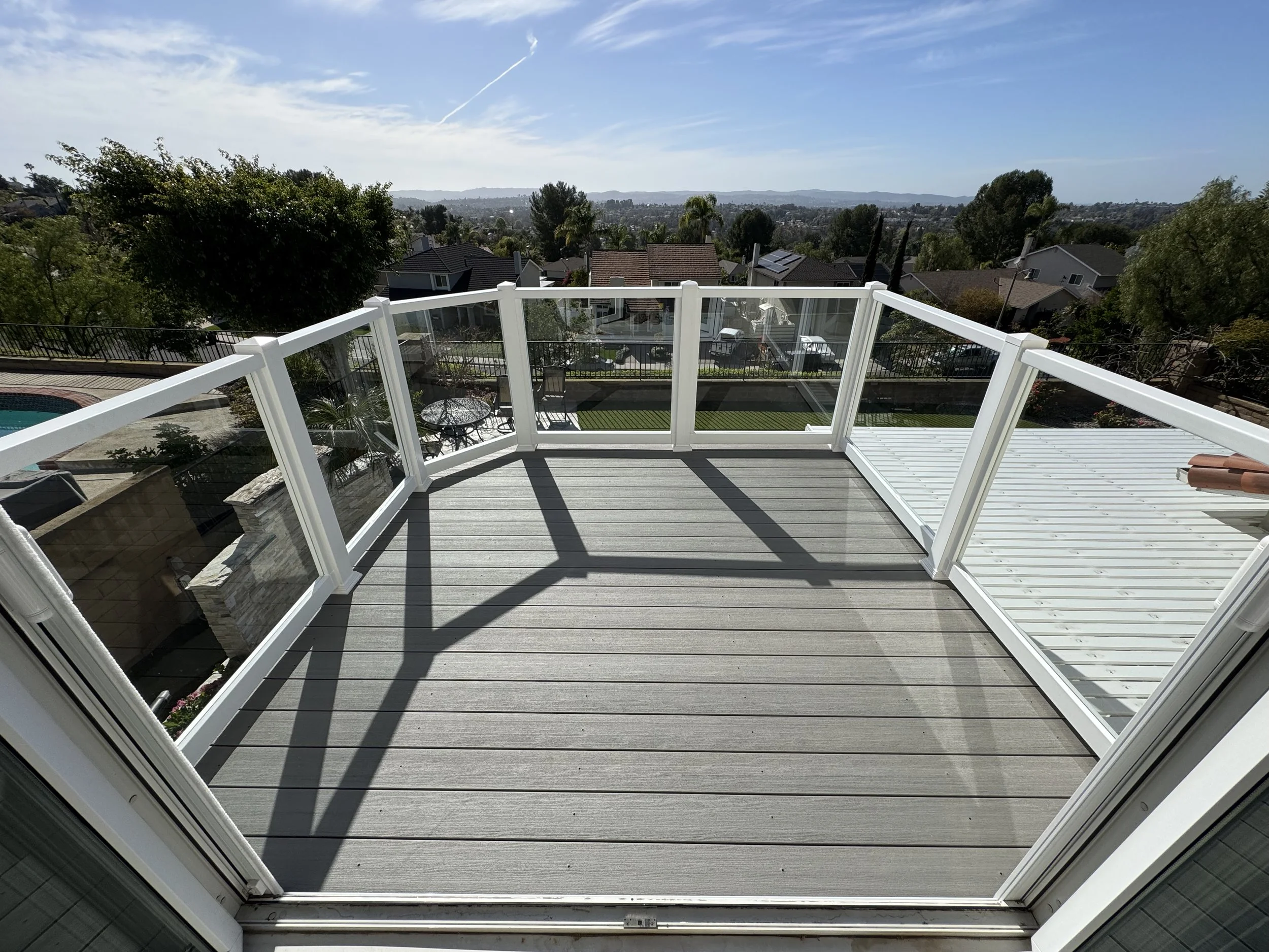 Fullerton balcony view with gray wood planks and white railing with glass panels overlooking a backyard with a pool, trees, houses, and a distant landscape under a partly cloudy sky, emphasizing custom vinyl patio covers and fences.