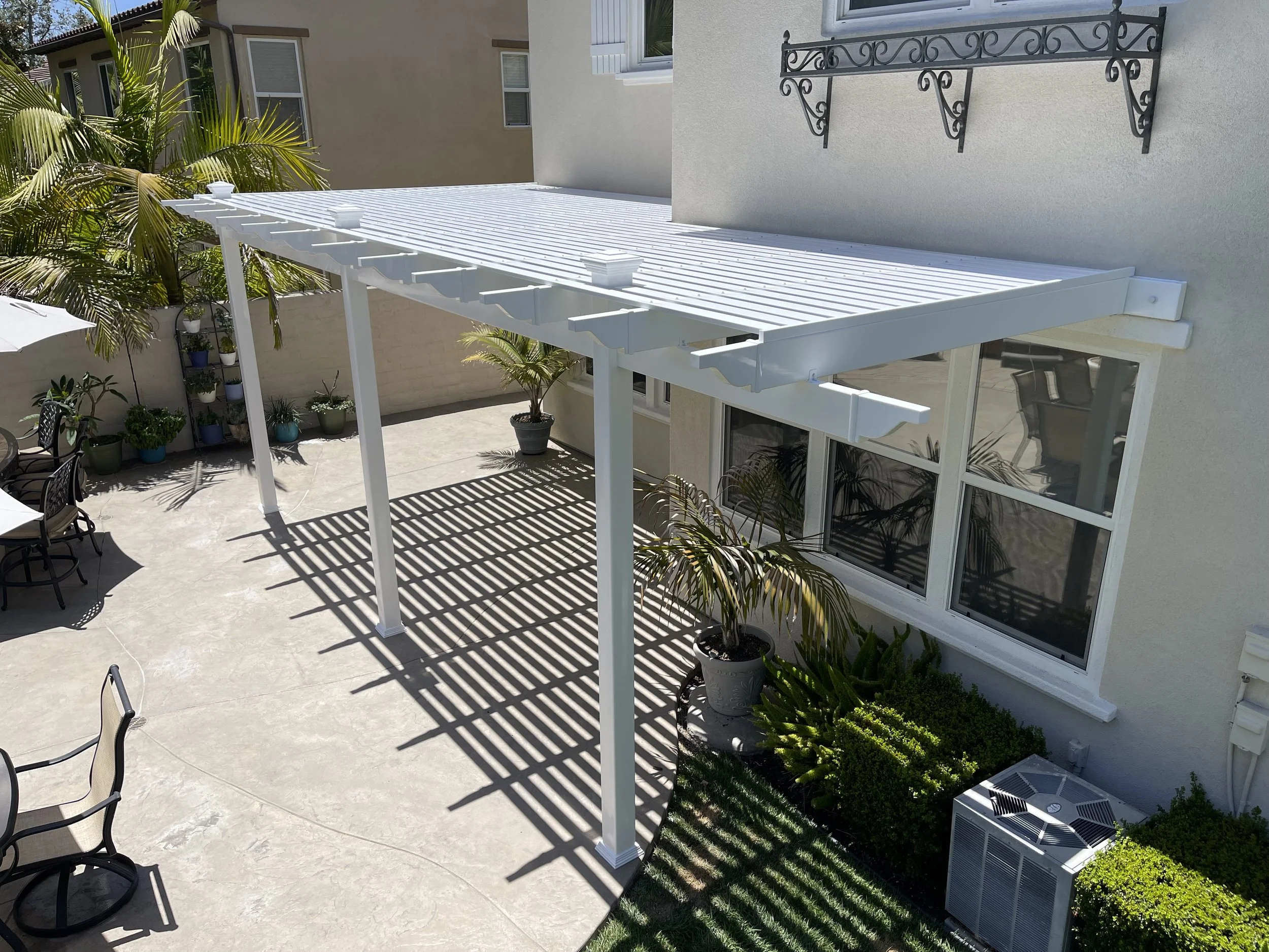 Backyard patio with a white metal pergola, potted plants, and patio furniture, under a sunny sky.