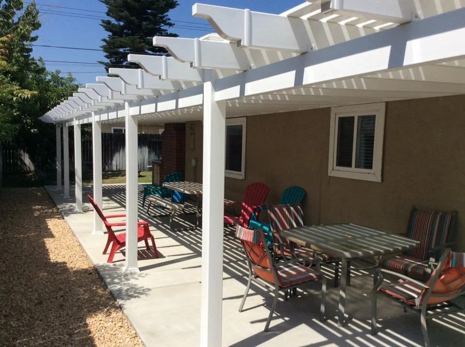 Brea backyard patio with a white vinyl pergola, chairs and tables, and a brick barbecue grill under bright sunlight, showcasing durable vinyl patio covers and custom gates in Orange County.
