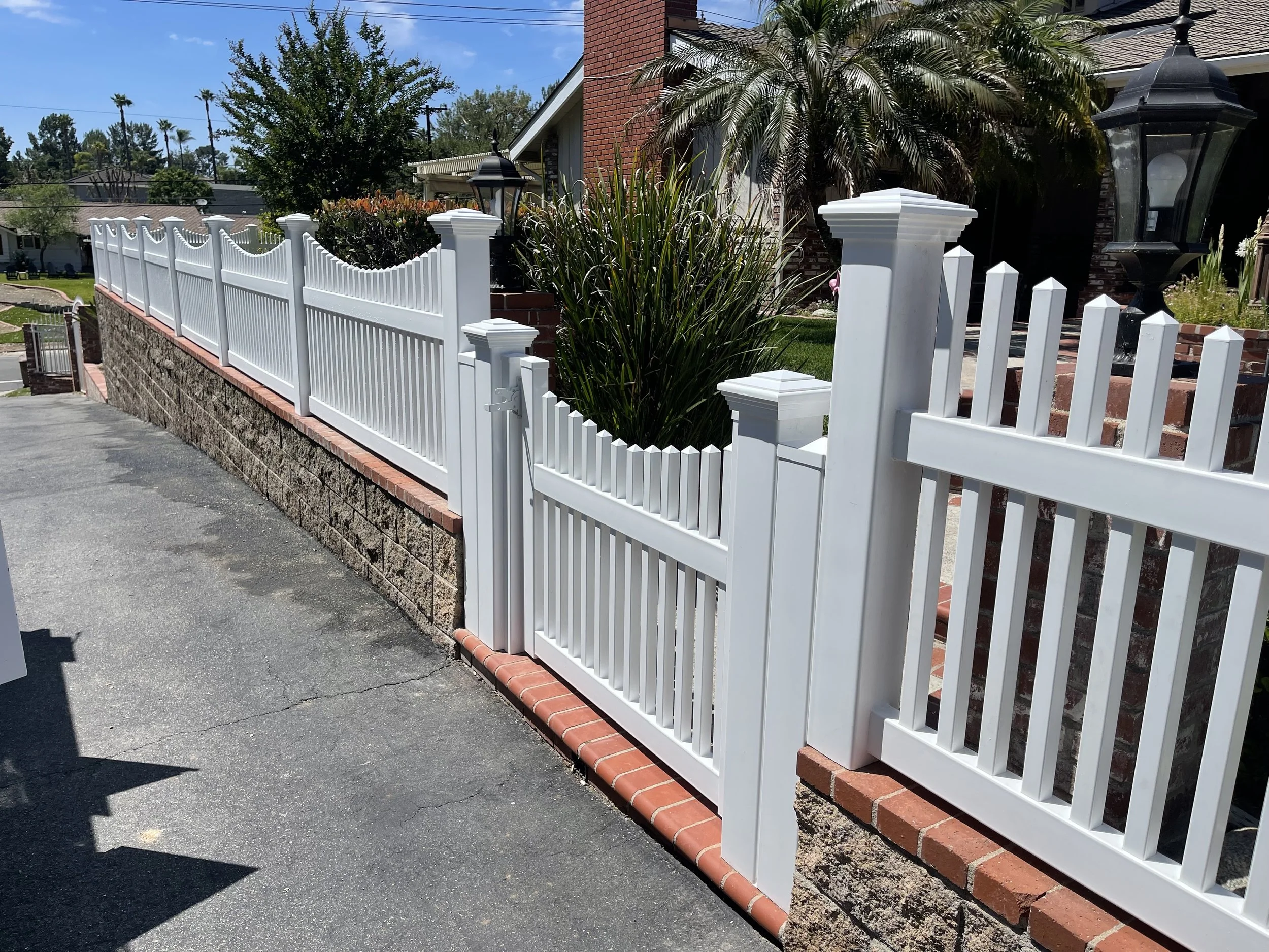Yorba Linda white vinyl picket fence with scalloped top and decorative posts, plus matching pedestrian gate on a brick-capped retaining wall along a driveway.
