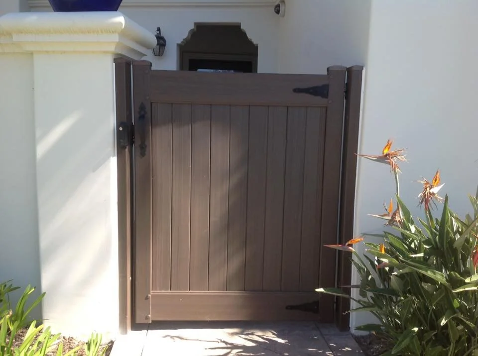 Dana Point brown vinyl privacy gate with black hardware, framed between stucco walls and accented by bird of paradise plants, stylish, durable, and low-maintenance fencing solution for coastal Orange County homes.