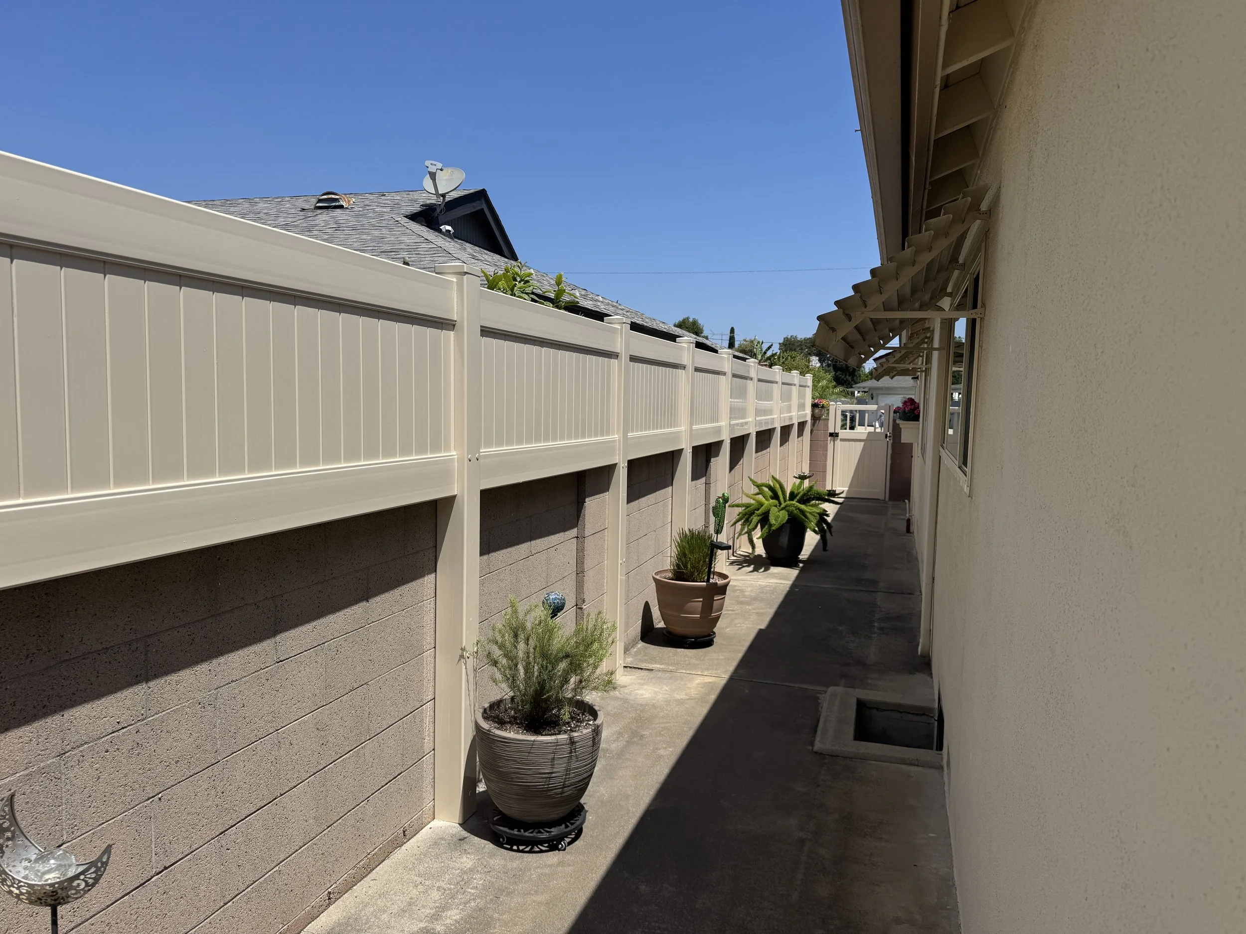 Buena Park side yard with potted plants along concrete pathway beside beige vinyl fence and stucco house wall with windows, showcasing durable vinyl fences and clean landscaping under clear blue sky.