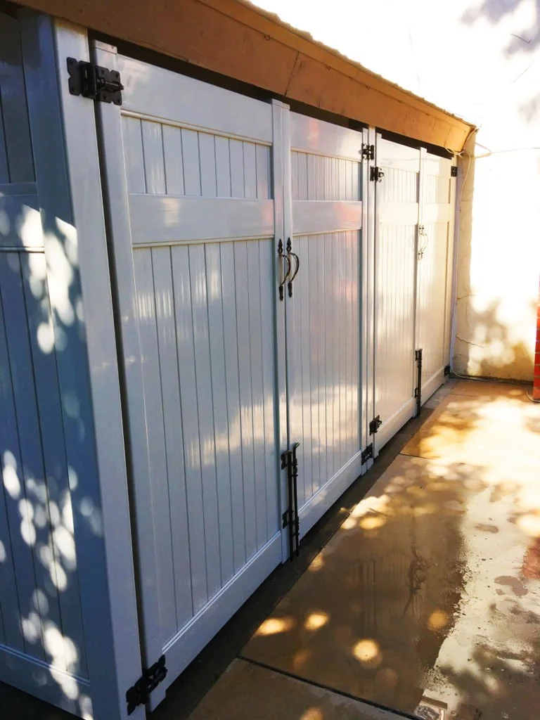 Fullerton white outdoor storage shed with double vinyl doors featuring black hinges and handles, situated on a concrete patio with tree shadows, illustrating how vinyl storage sheds complement fencing and patio cover projects in Orange County.