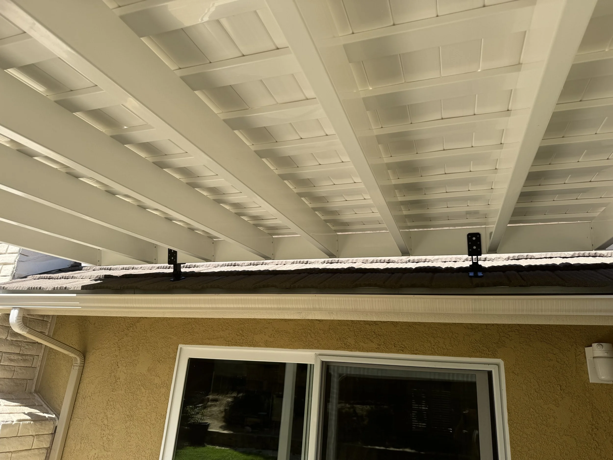 Looking up at a porch with a white wooden ceiling, beige stucco wall, window, and a small black metal bracket supporting the roof.