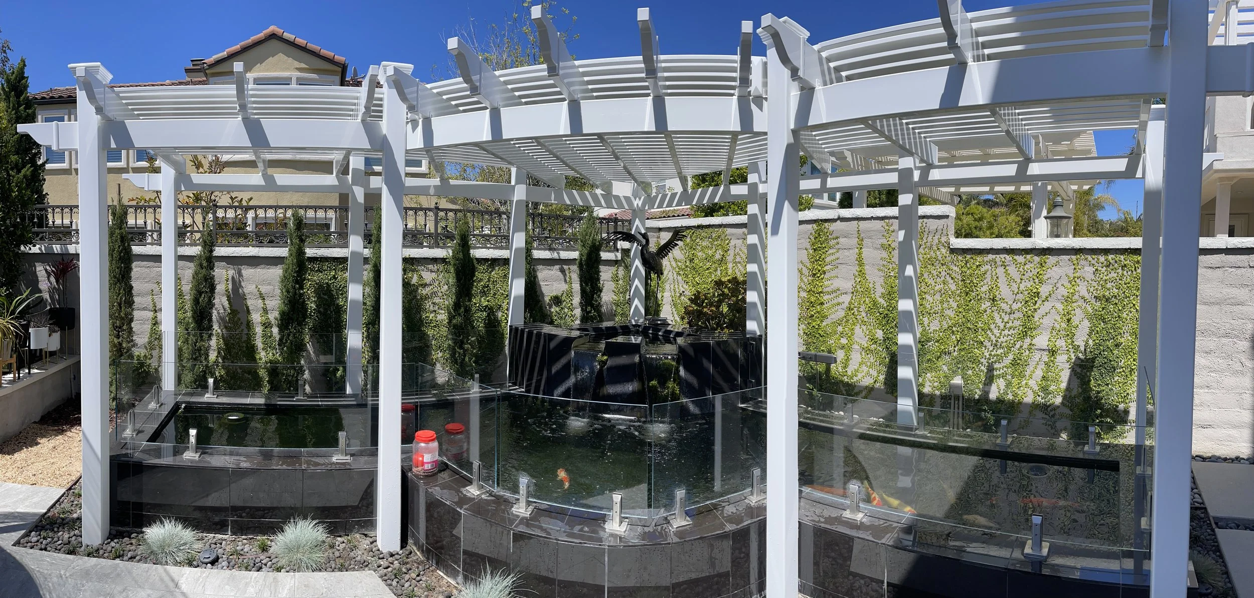 Backyard with white pergola, small pond with koi fish, decorative waterfall, and lush green plants on a sunny day.