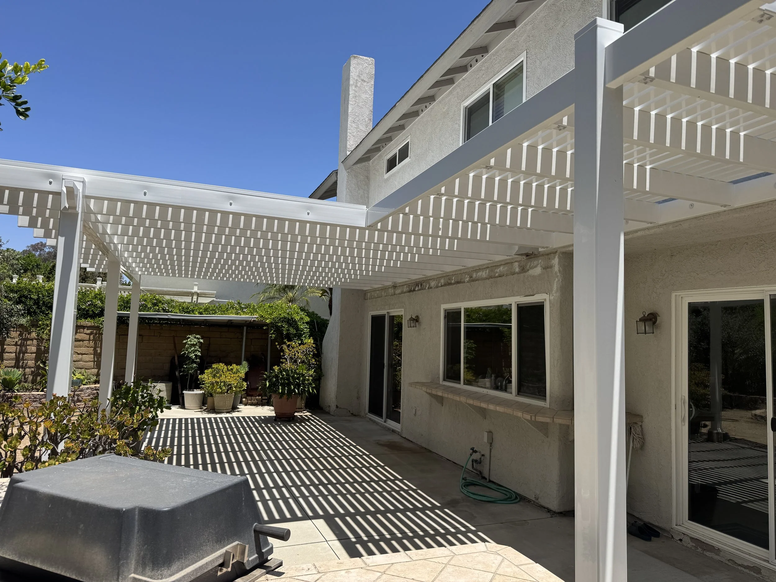 Rancho Santa Margarita backyard patio area with a white vinyl slatted pergola casting shadows, potted plants against a brick fence, and a barbecue grill in the foreground beside a two-story house with sliding glass doors and windows.