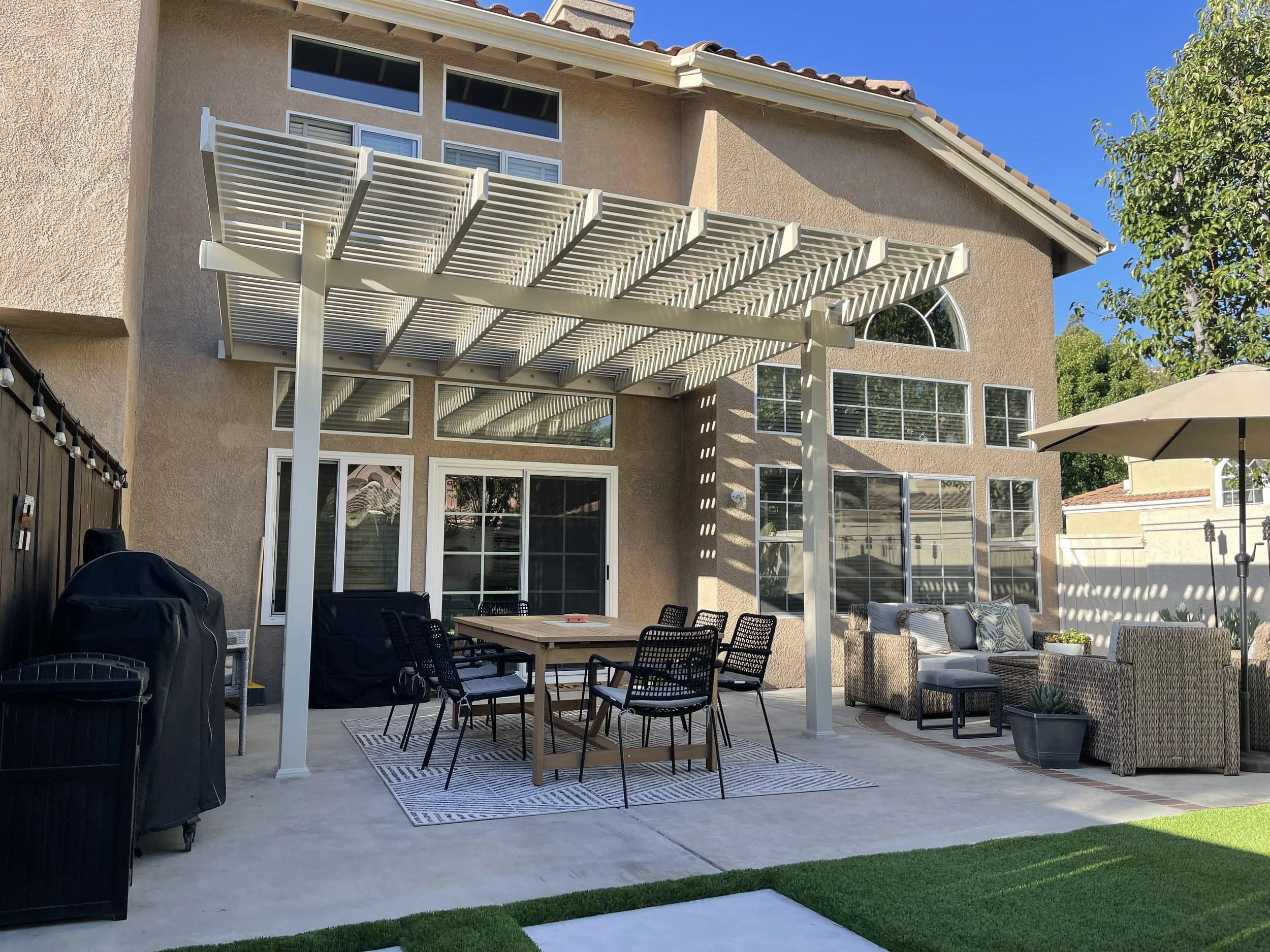 Backyard patio with outdoor furniture, a table with eight chairs, a large beige umbrella, a lounge sitting area with a sofa and armchairs, a table with potted plants, and a white pergola structure, with a two-story house with large windows in the bac