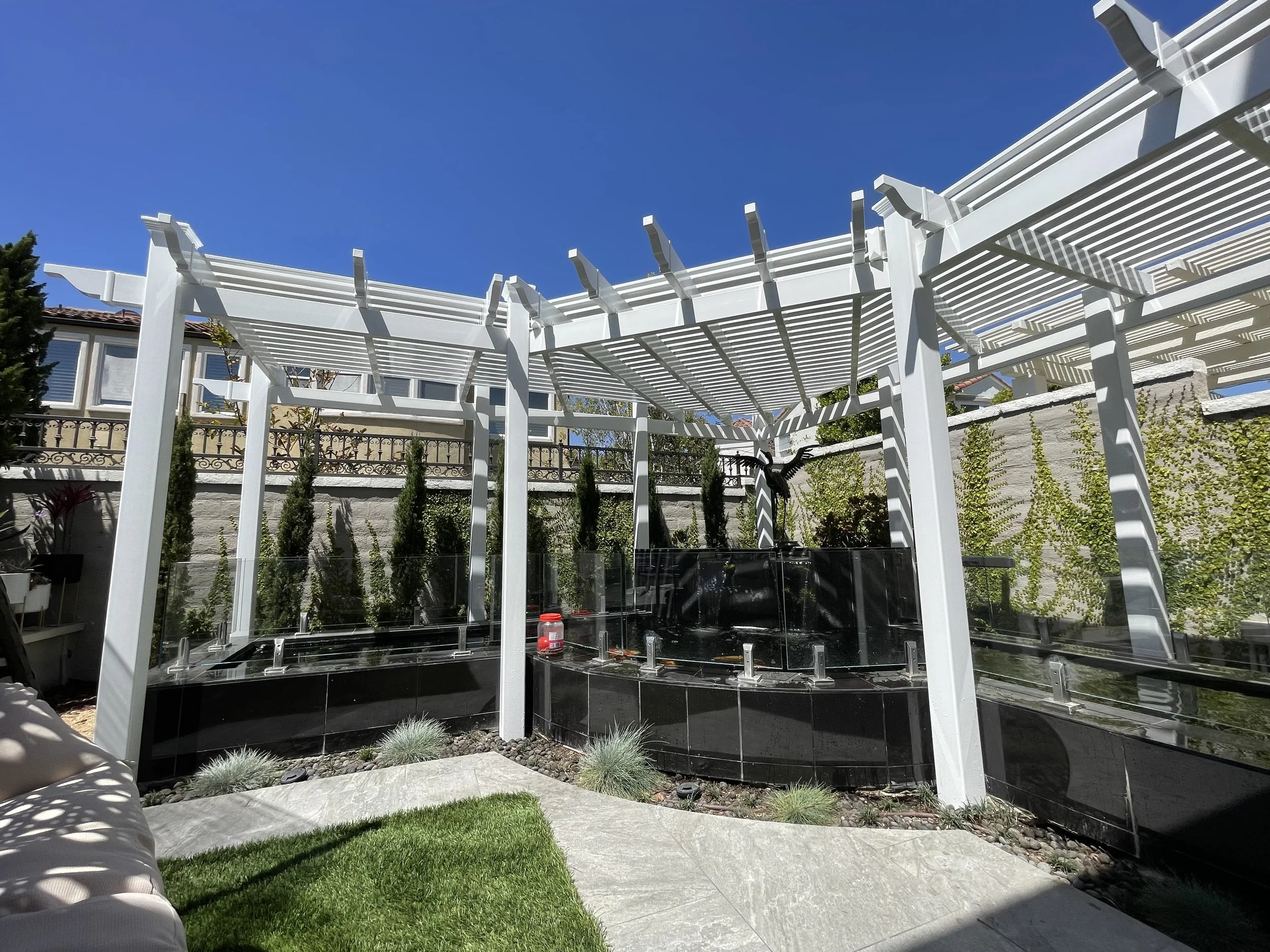 Backyard patio with a white pergola and black water feature, surrounded by a stone fence covered with green vines and tall, narrow trees.