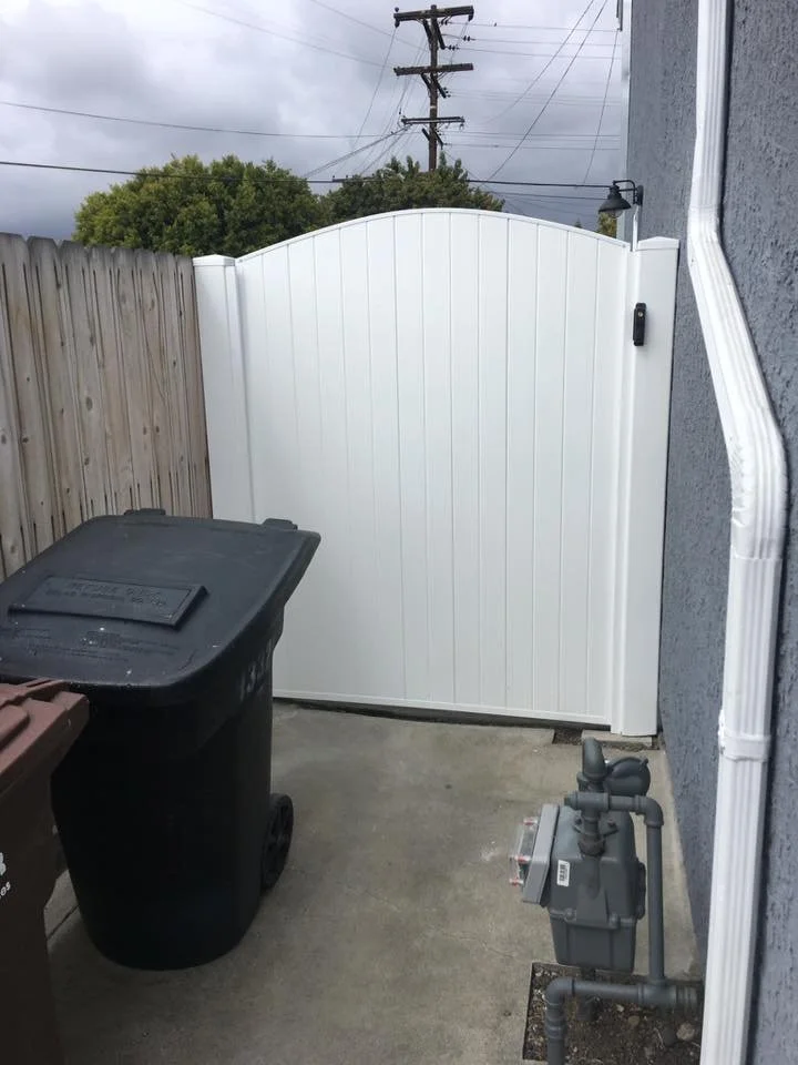 Garden Grove small outdoor area with white vinyl privacy fence enclosing a utility space with trash and recycling bins and utility meters near a gray house wall, with power lines and trees in the background, showcasing durable vinyl fencing for side