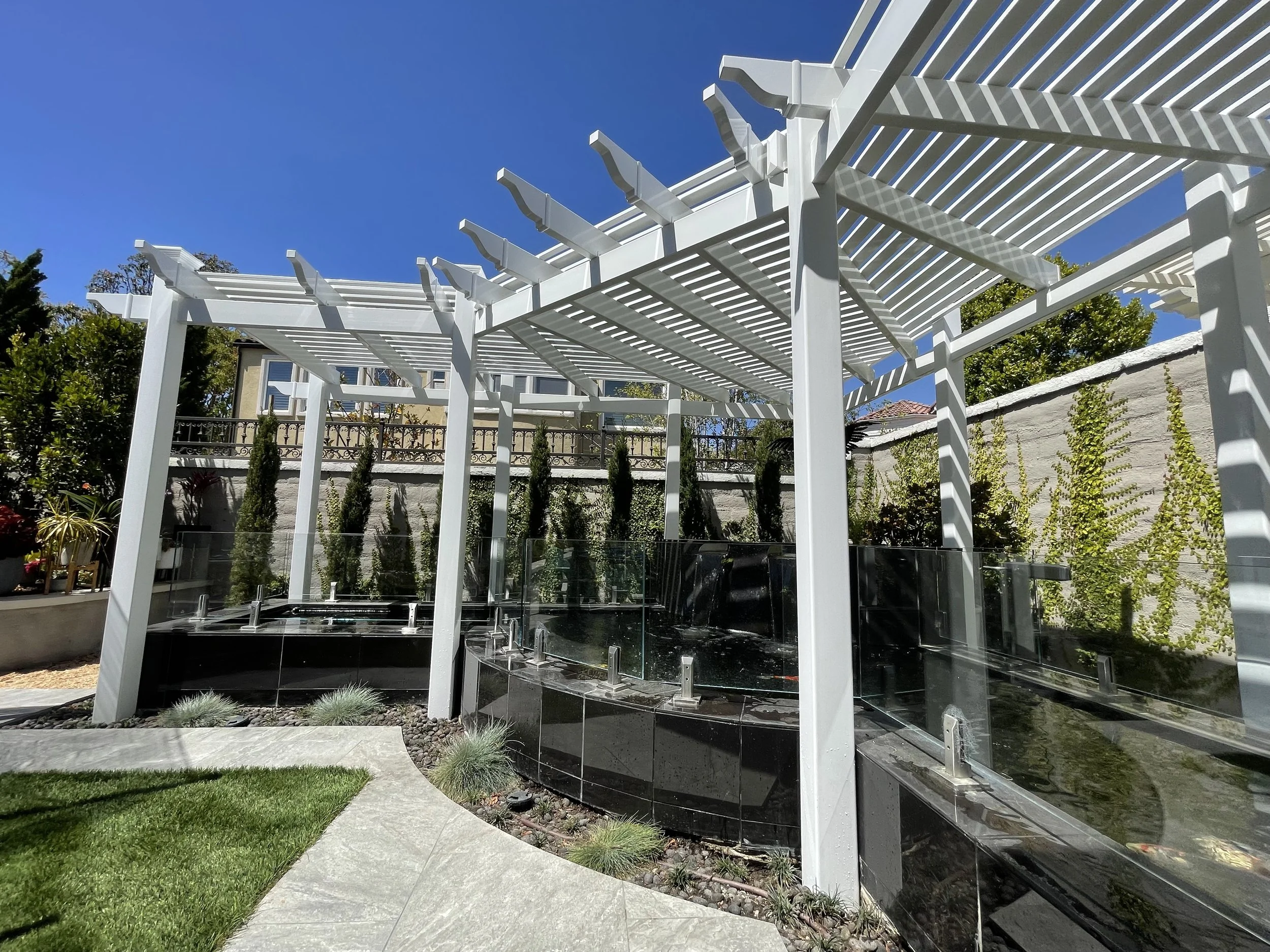 A modern backyard with a white pergola, glass fencing, green grass, and plants against a bright blue sky.