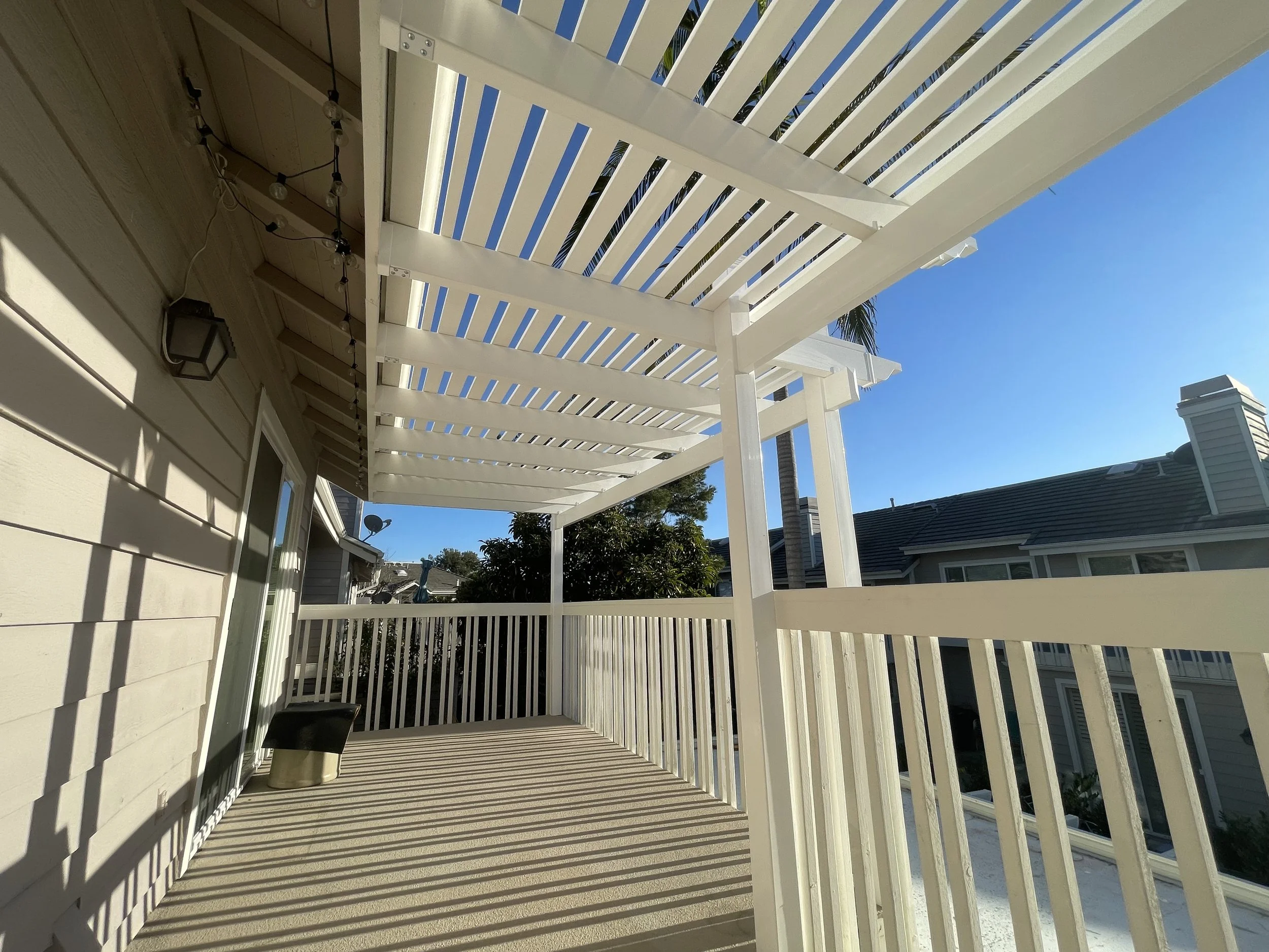 Upward view of a balcony with white wooden railings and a pergola casting patterned shadows, adjacent to a beige house with a door, lantern-style light, string lights, and a small black and silver trash can, under clear blue sky.