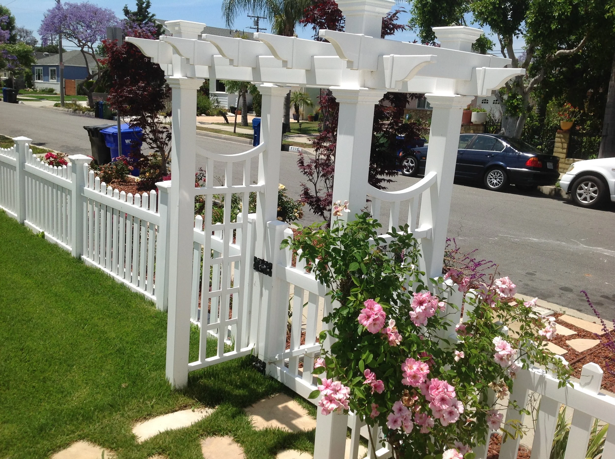 Garden Grove white vinyl picket fence with matching arbor gate, decorative lattice design, and blooming pink climbing roses, charming entryway enhancing curb appeal for Orange County homes.
