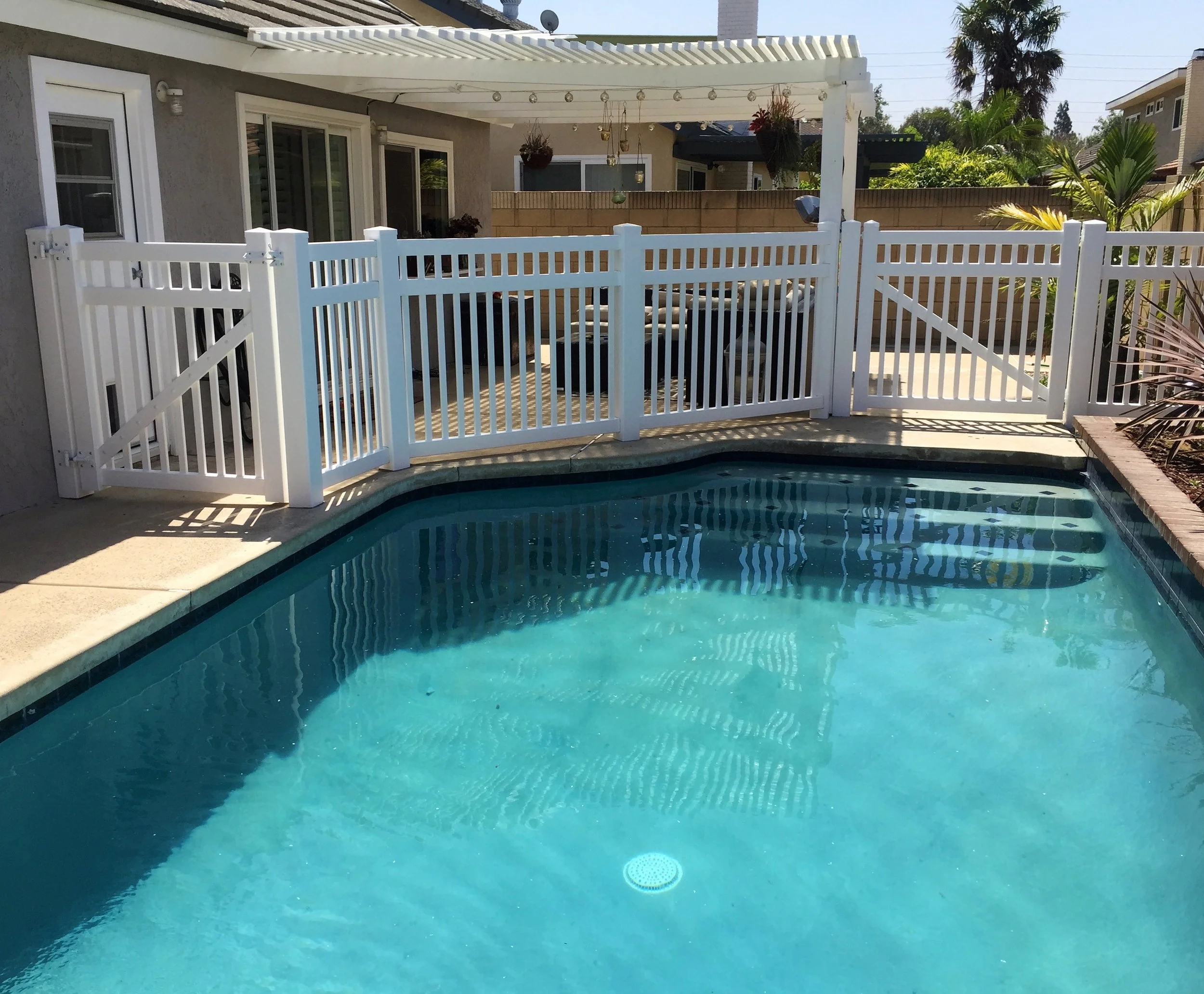 Cypress residential backyard with small swimming pool, white vinyl safety fence, patio furniture and potted plants around the pool with palm trees and neighboring houses in Orange County.