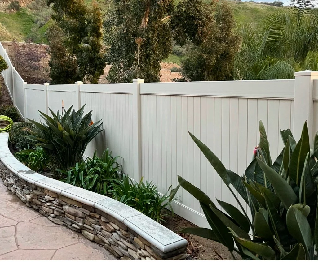 Costa Mesa white vinyl privacy fence with decorative panels surrounded by green plants and trees and a stone-bordered flower bed in the foreground, showcasing premium vinyl fencing solutions.