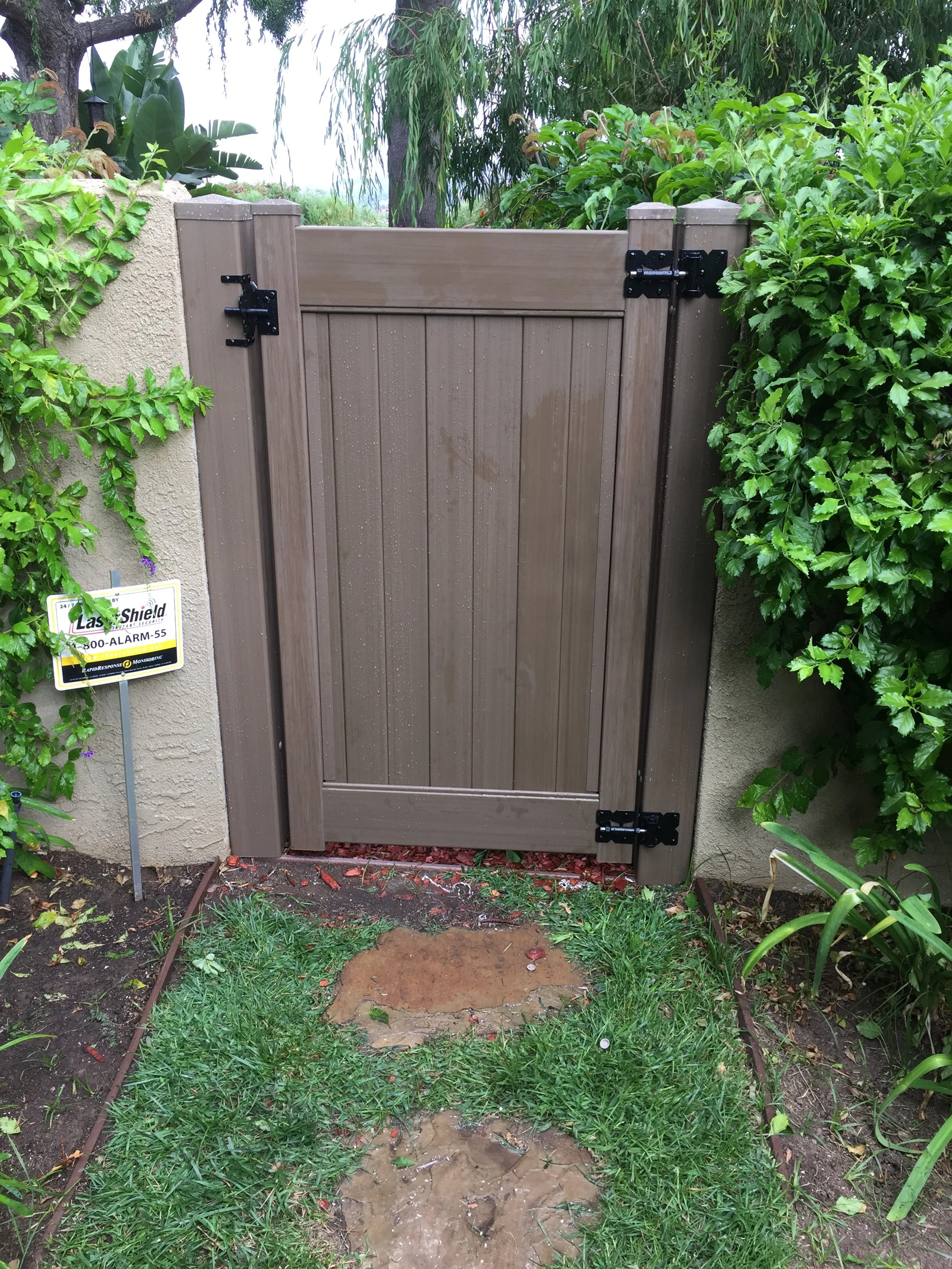 Laguna Niguel brown vinyl side gate with black hinges between stucco pillars and dense hedges, stepping-stone path on lawn, durable, low-maintenance backyard fencing.