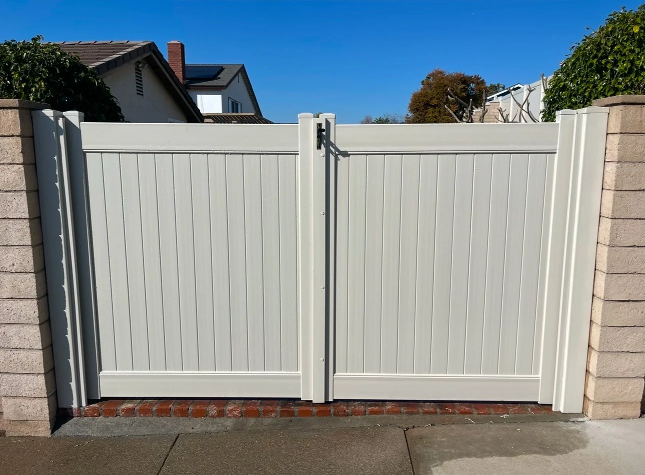 Laguna Woods white vinyl double gate with vertical panels and decorative top rails set between brick pillars on a concrete sidewalk, with a clear blue sky in the background, showcasing premium vinyl gates and fencing.