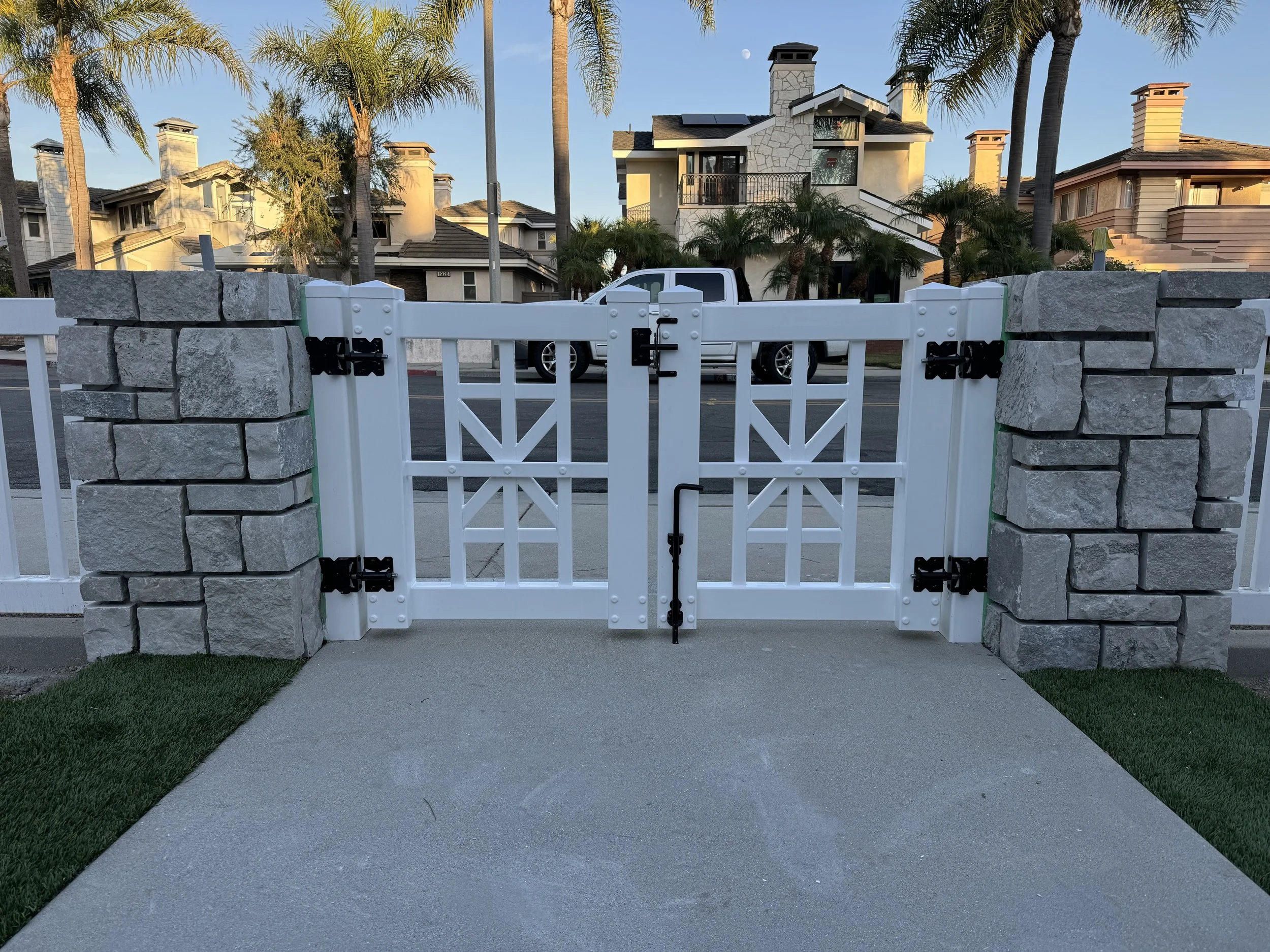 Los Alamitos vinyl gate between gray stone pillars with black hinges and latch, on concrete walkway flanked by lawns and palm-lined street, showcasing custom vinyl gates and fencing in Orange County.