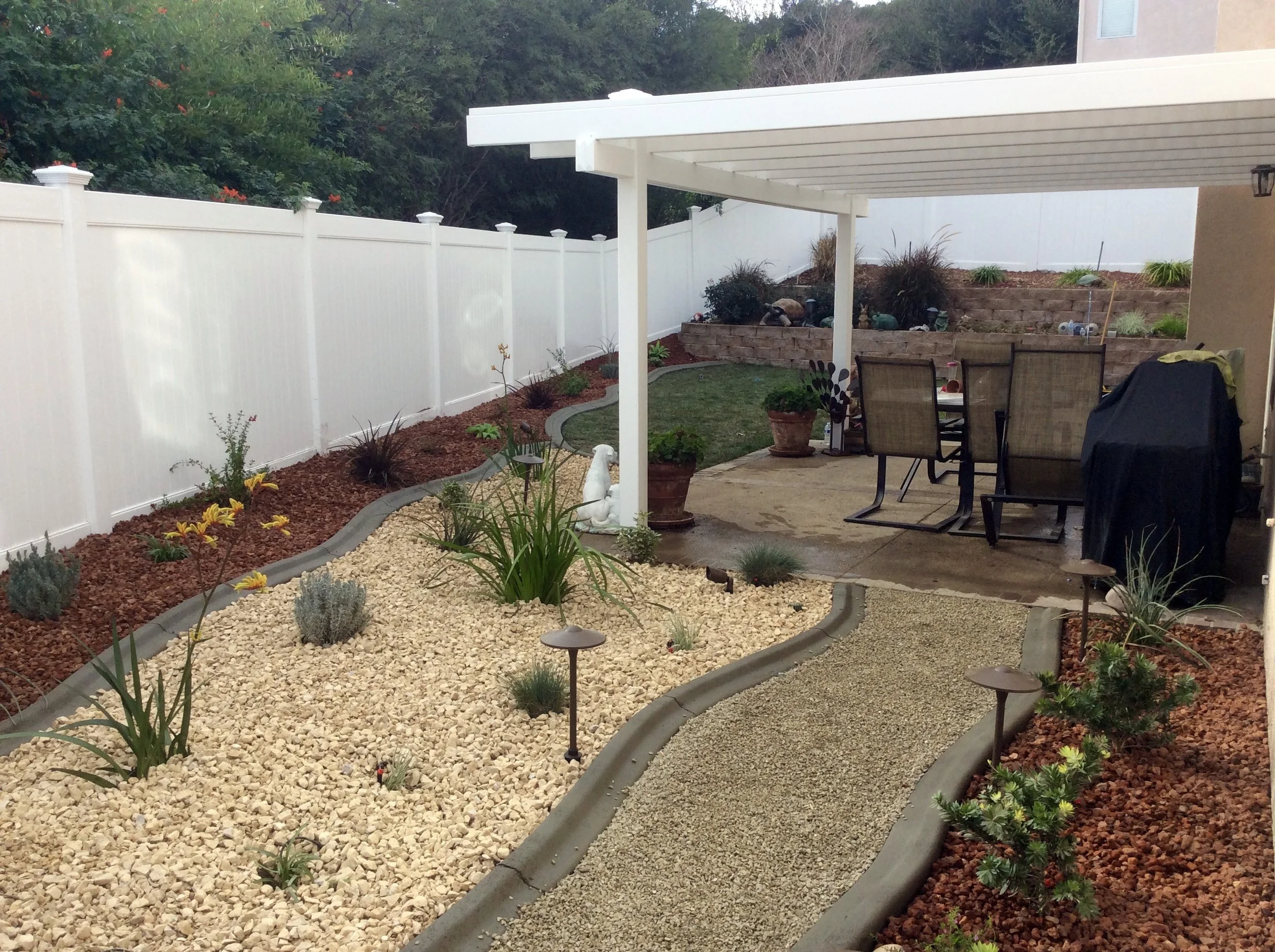 Laguna Woods backyard patio with a white vinyl fence, a covered dining area with chairs, a gravel pathway, and landscaped garden beds with plants and decorative rocks, illustrating premium vinyl fencing, gates, and patio covers in Orange County.