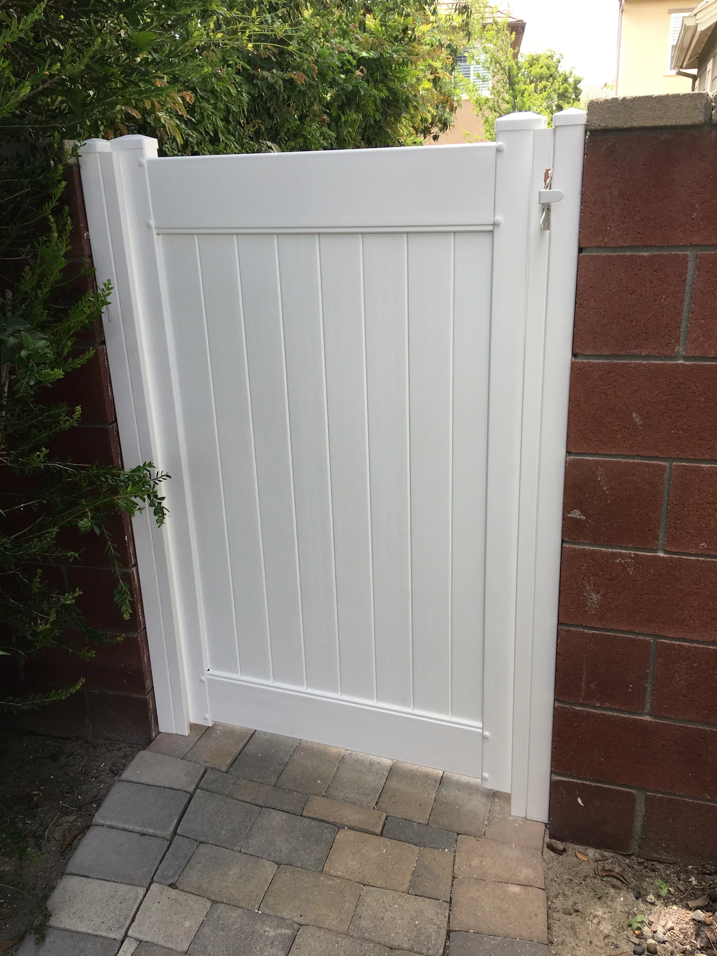 Westminster white vinyl privacy gate with vertical panels and secure latch, framed between red brick walls and paver walkway, low-maintenance, durable fencing option for Orange County homes.