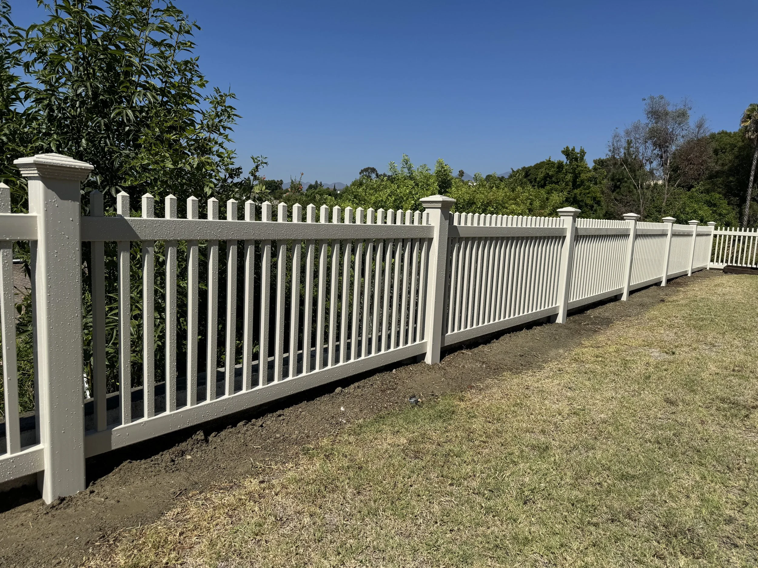 San Clemente white picket fence running along a grassy yard with trees under a clear blue sky, highlighting quality vinyl fencing and custom gates.