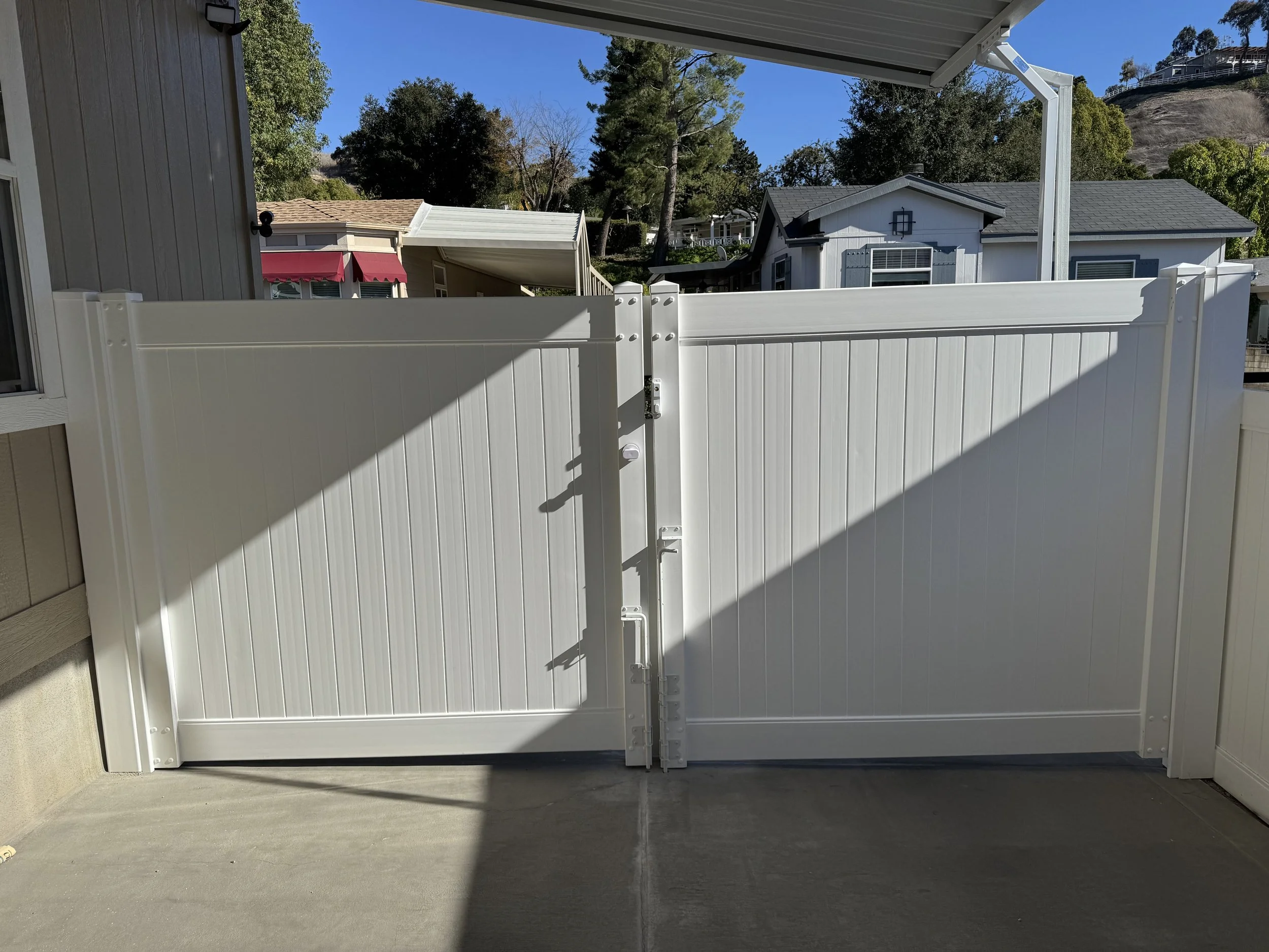 Stanton white vinyl fence gate on a concrete patio with clean lines and decorative posts, with trees and houses in the background under a clear blue sky, showcasing durable vinyl fencing and gates.
