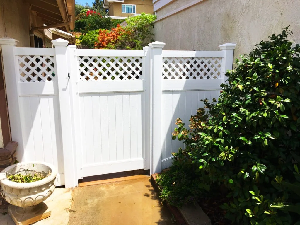 Costa Mesa white vinyl privacy fence gate with lattice top next to lush green shrubbery and a decorative garden planter in a backyard area, highlighting durable vinyl fencing and patio covers for Orange County homes.
