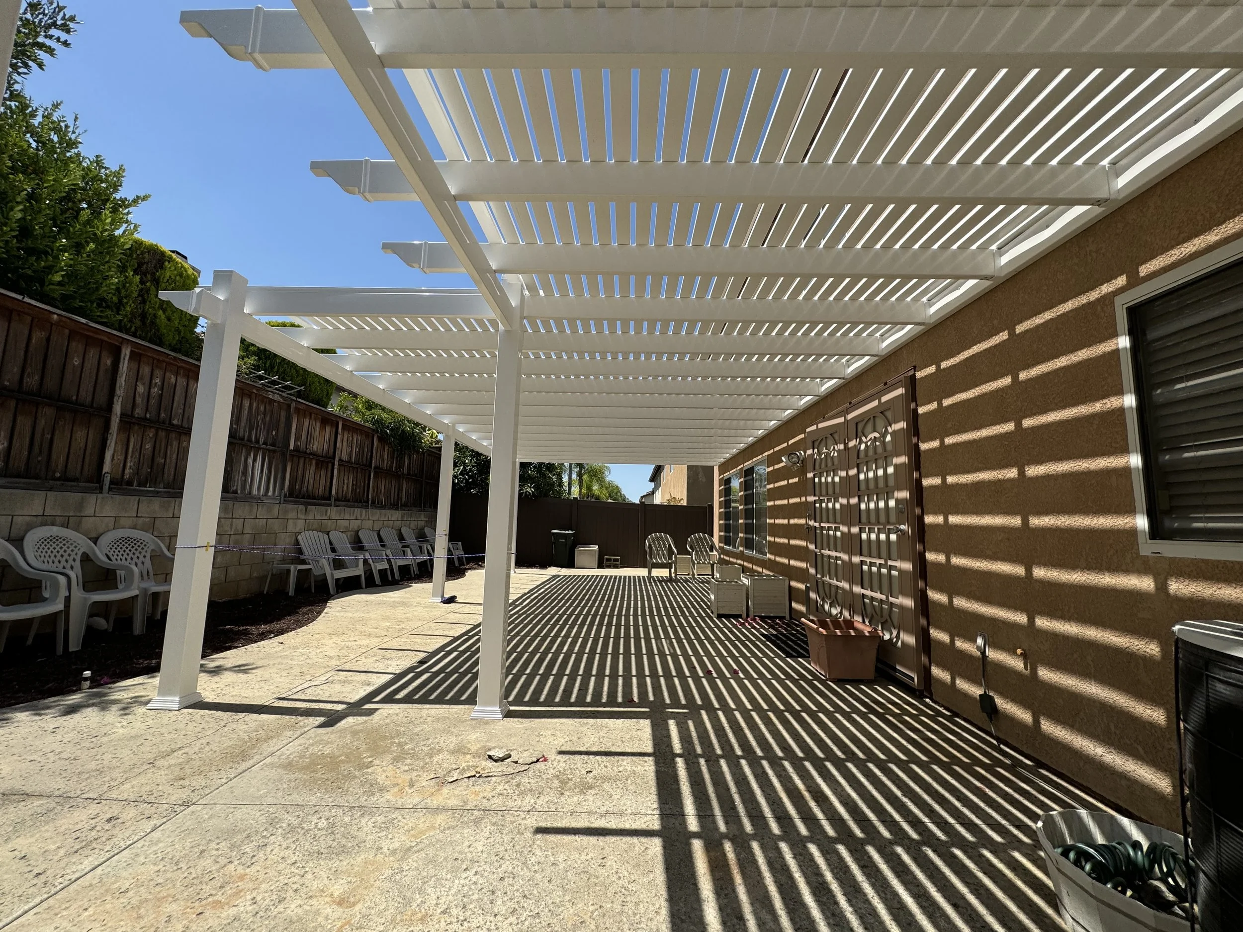 Backyard patio with white pergola casting striped shadows on the concrete floor. Outdoor chairs along the fence, and potted plants near the house. The house has a tan stucco wall with windows and a door. Clear blue sky overhead.