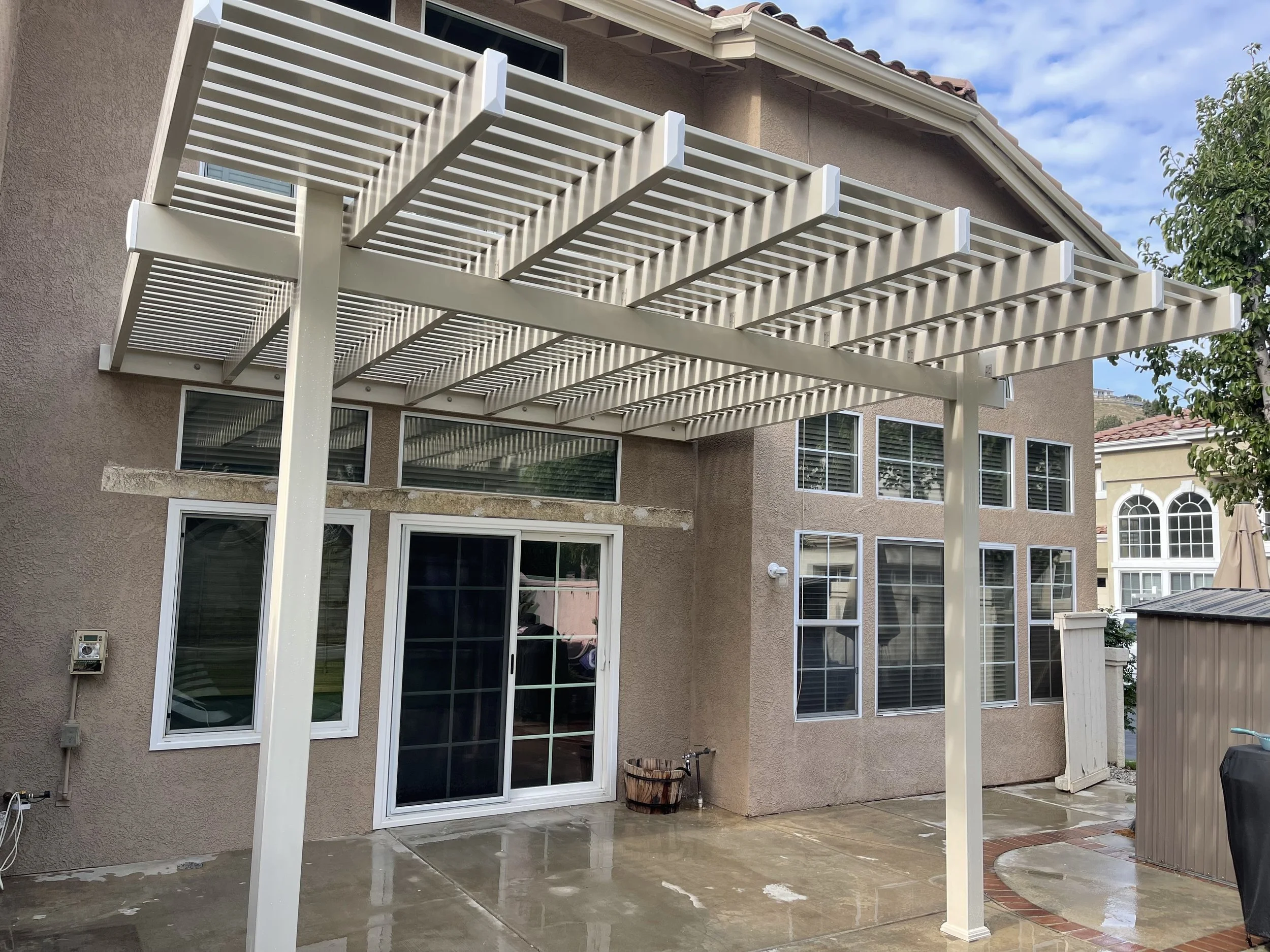 White pergola over a wet concrete backyard patio with a beige house, sliding glass door, and multiple windows.