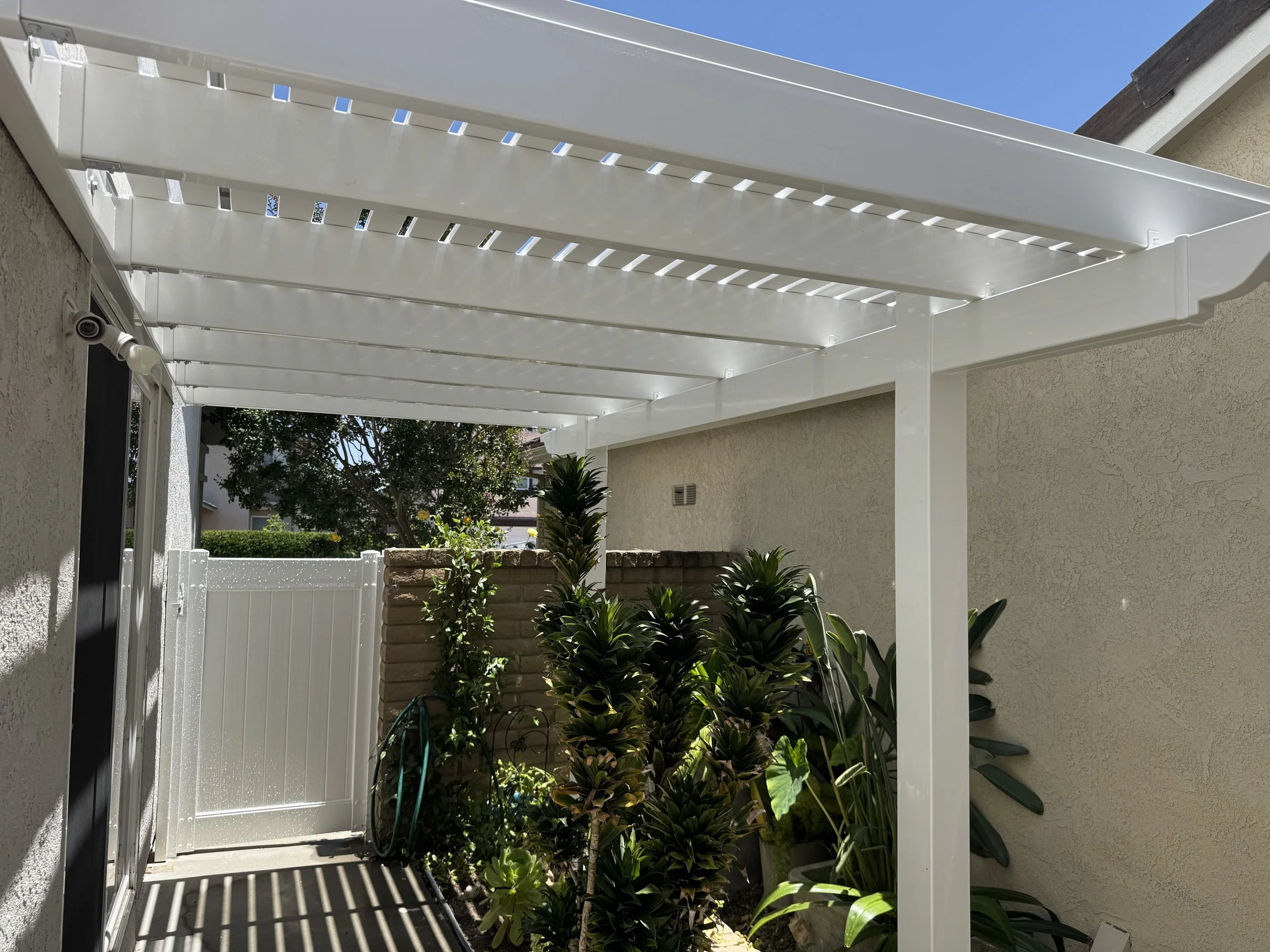 Lake Forest white vinyl pergola attached to a house, shading a small outdoor area with plants and a garden hose under a sunny sky, demonstrating durable vinyl patio covers and fencing services in Orange County.