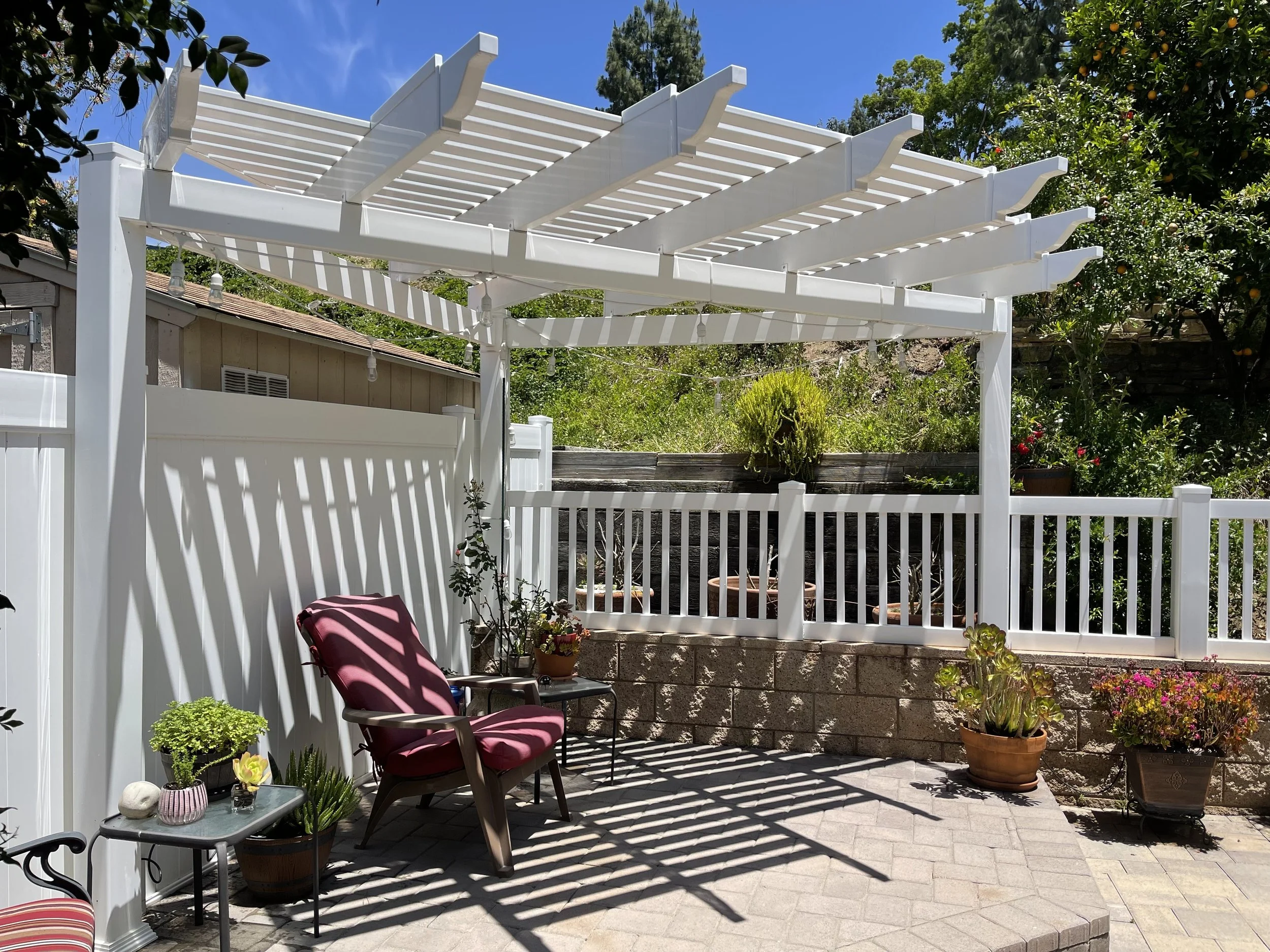 A backyard patio with a white wooden pergola casting striped shadows, a maroon cushioned chair, potted plants, and a white fence, during a sunny day.