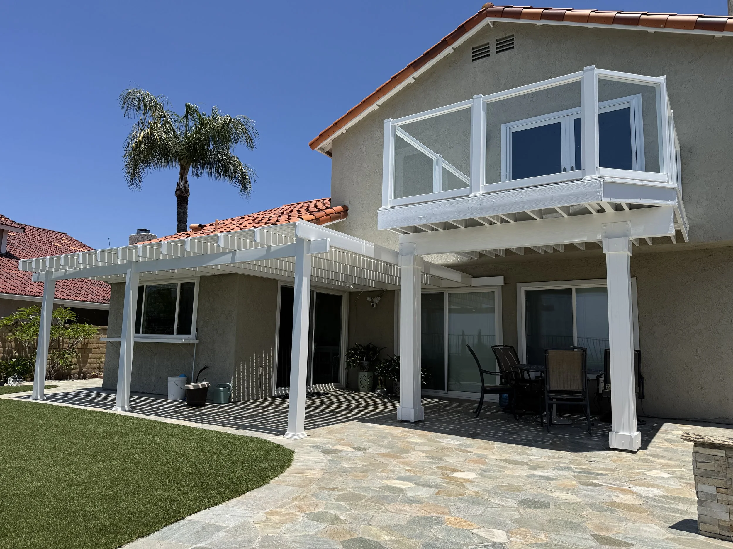 Backyard patio with a white pergola and an elevated balcony with glass railings on a two-story house. There is a stone pathway and a patch of green grass with a palm tree in the background.